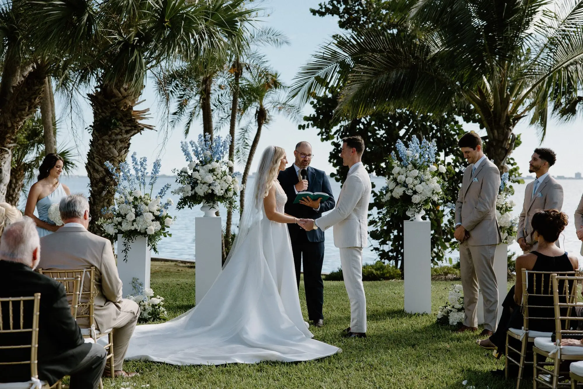 Bride and Groom Wedding Ceremony Vow Exchange Wedding Portrait | White Roses and Florals with Blue Delphiniums in White Vases on White Pedestals Wedding Ceremony Floral Arrangement Decor Ideas | Ocean View Wedding Reception Inspiration | Gold Chiavari Ceremony Chairs Ideas | Sarasota Florist Beneva Flowers