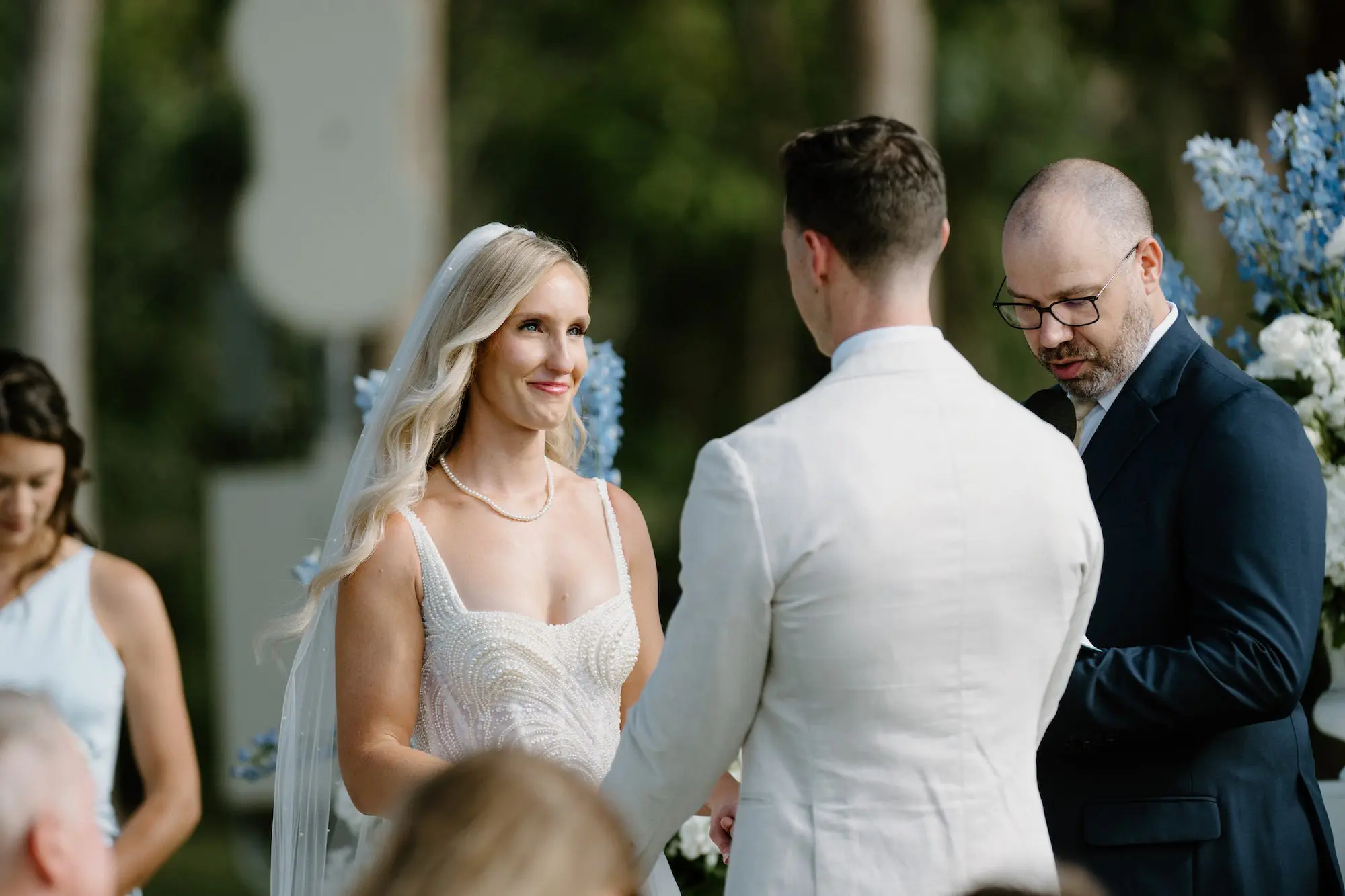 Bride and Groom Wedding Ceremony Vow Exchange Wedding Portrait | White Roses and Florals with Blue Delphiniums in White Vases on White Pedestals Wedding Ceremony Floral Arrangement Decor Ideas | Ocean View Wedding Reception Inspiration | Gold Chiavari Ceremony Chairs Ideas | Sarasota Florist Beneva Flowers