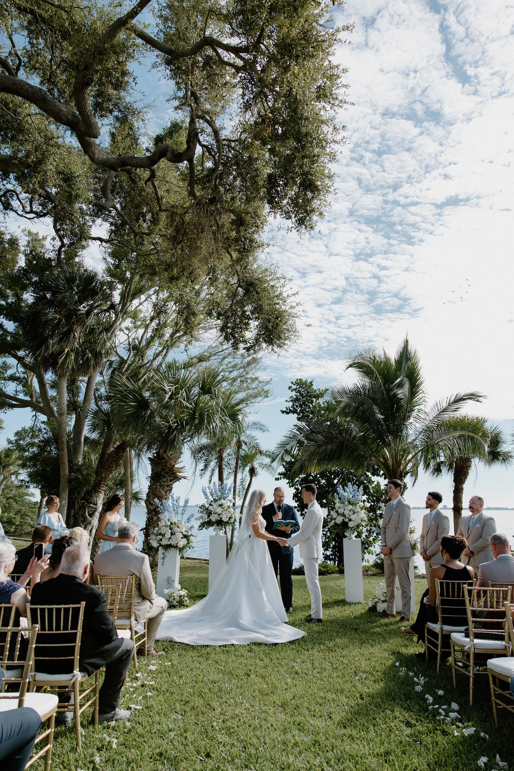 Bride and Groom Wedding Ceremony Vow Exchange Wedding Portrait | Oceanview Timeless Wedding Reception Venue Inspiration | White Roses and Florals with Blue Delphiniums in White Vases on White Pedestals Wedding Ceremony Floral Arrangement Decor Ideas | Ocean View Wedding Reception Inspiration | Gold Chiavari Ceremony Chairs Ideas | Sarasota Florist Beneva Flowers