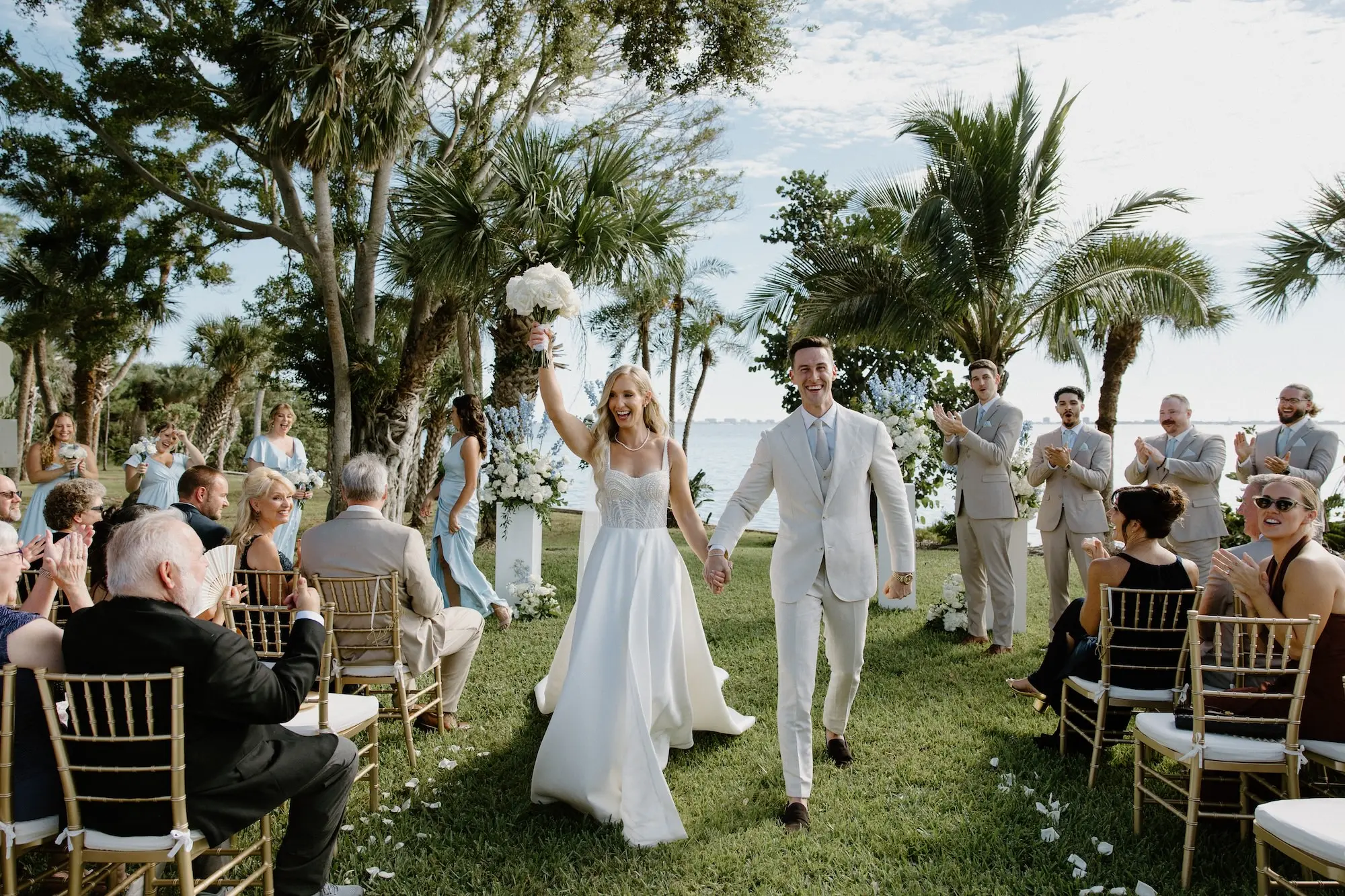 Bride and Groom Just Married Wedding Portrait | White Roses and Florals with Blue Delphiniums in White Vases on White Pedestals Wedding Ceremony Floral Arrangement Decor Ideas | Ocean View Wedding Reception Inspiration | Gold Chiavari Ceremony Chairs Ideas | Sarasota Florist Beneva Flowers