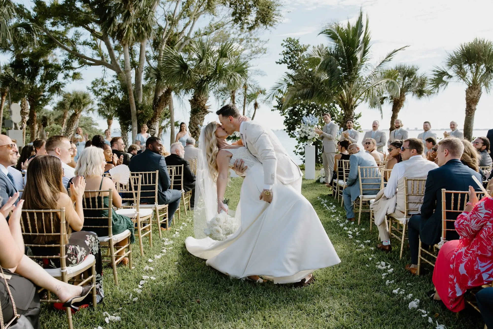 Bride and Groom Just Married Kiss Wedding Portrait | White Roses and Florals with Blue Delphiniums in White Vases on White Pedestals Wedding Ceremony Floral Arrangement Decor Ideas | Ocean View Wedding Reception Inspiration | Gold Chiavari Ceremony Chairs Ideas | Sarasota Florist Beneva Flowers