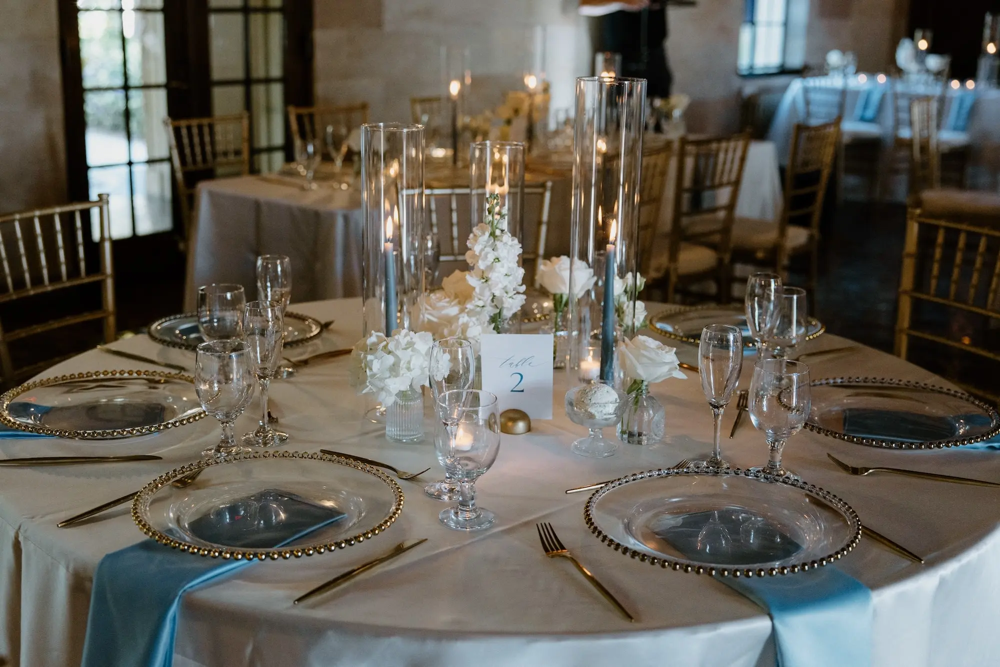 Classic Ballroom Wedding Reception | Modern Gold Chiavari Chairs | Bud Vase Centerpiece White Roses and Orchid Flower Arrangement Ideas | Clear Chargers with Gold Rims and Satin Light Blue Napkins Wedding Reception Tablescape Decor Inspiration | Sarasota Florist Beneva Flowers | Venue Powel Crosely Estate