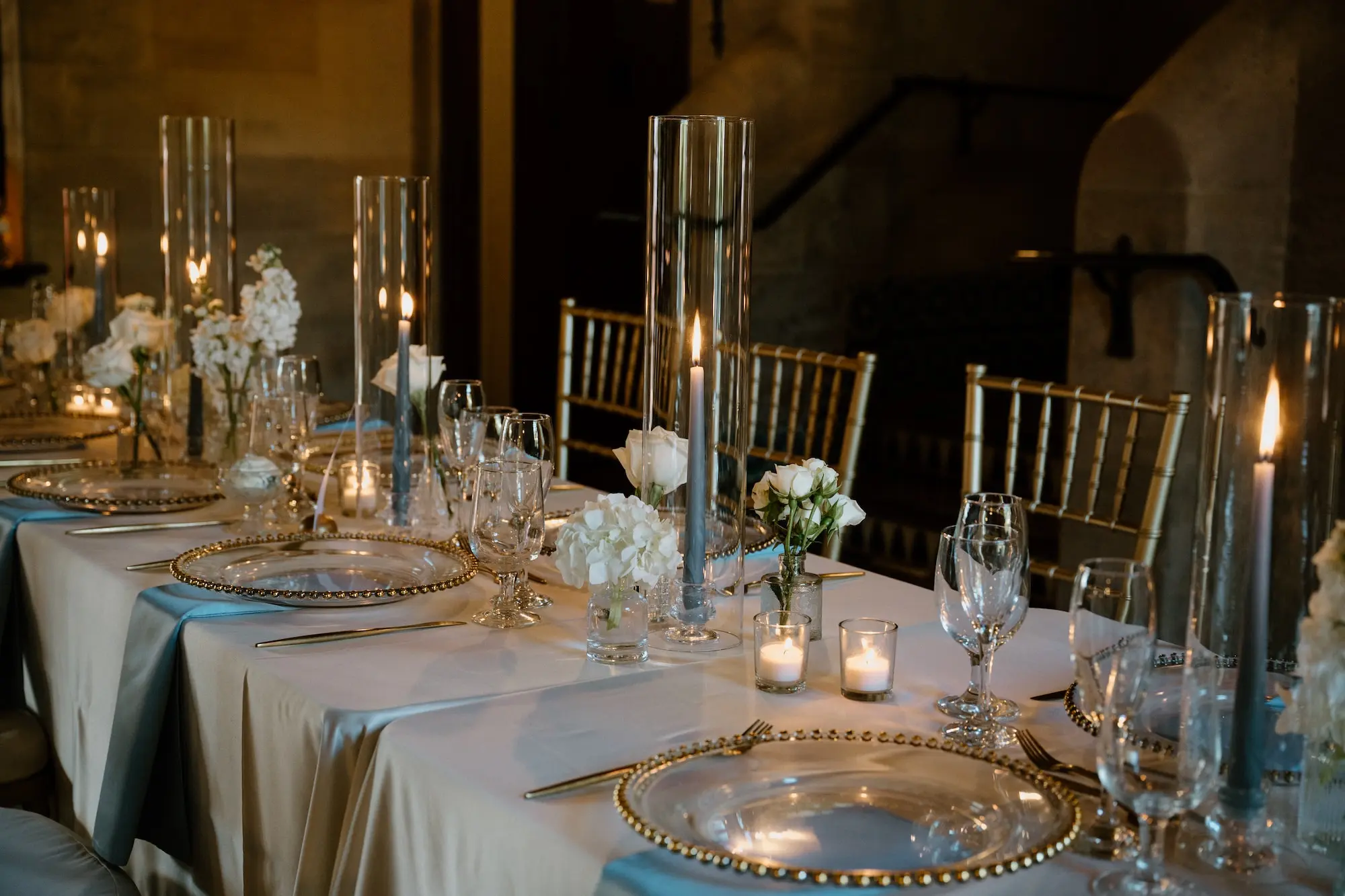 Classic Ballroom Wedding Reception | Bud Vase Centerpiece White Roses and Orchid Flower Arrangement Ideas | Gold Chiavari Reception Chairs | Blue Taper Candles in Glass Vases Wedding Reception Tablescape Decor Inspiration | Clear Chargers with Gold Rims on Blue Satin Napkins Reception Decor Ideas | Sarasota Florist Beneva Flowers