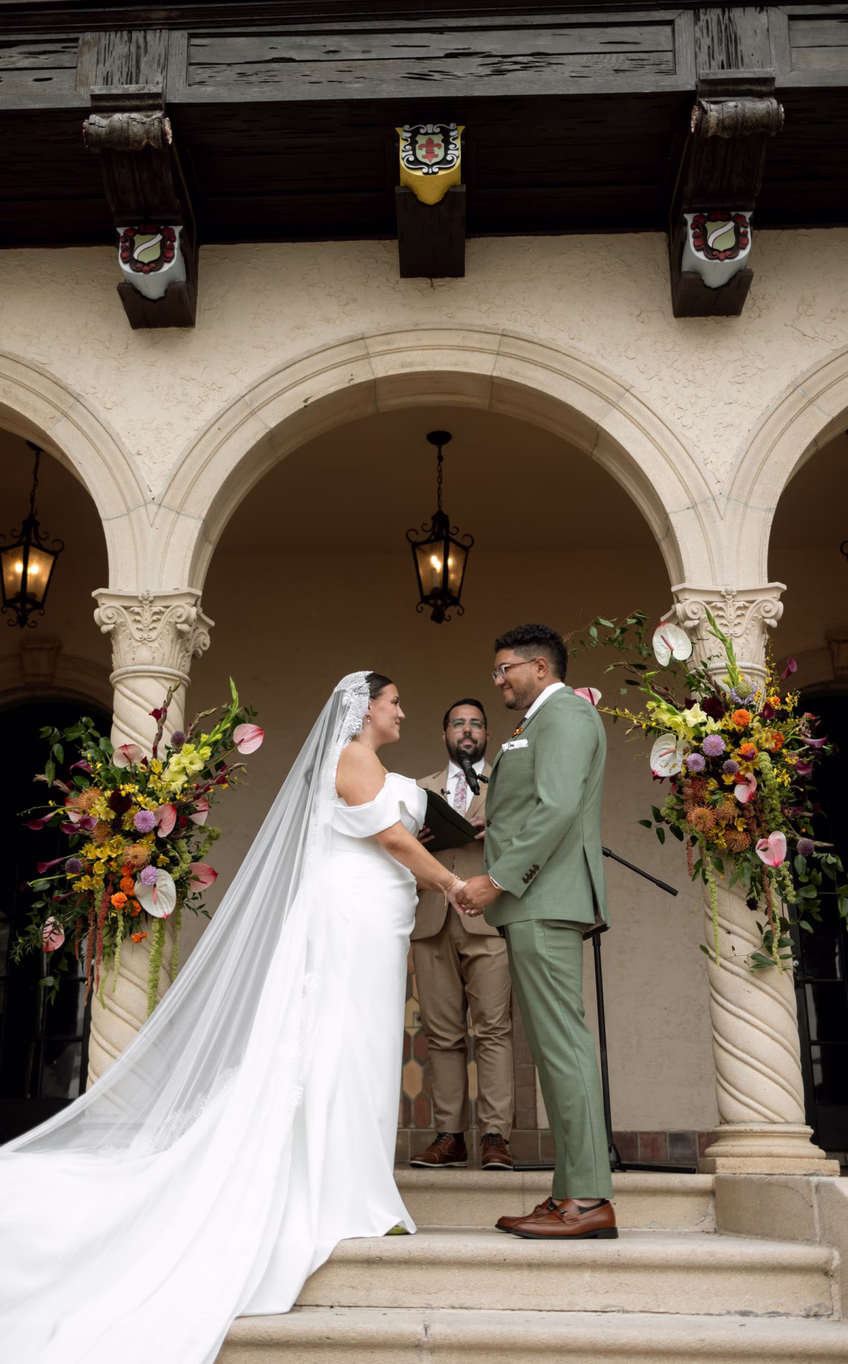 Bride and Groom Wedding Ceremony Vow Exchange Wedding Portrait | White Off the Shoulder Made with Love Wedding Gown Inspiration | Traditional Lace Cathedral Length Wedding Veil Ideas | Green Groom Suit Inspiration | Tropical Fall Colors Yellow, Orange, Purple, Pink, Red Florals with Greenery Wedding Ceremony Altar Decor Inspiration | Sarasota Event Venue Powel Crosley Estate
