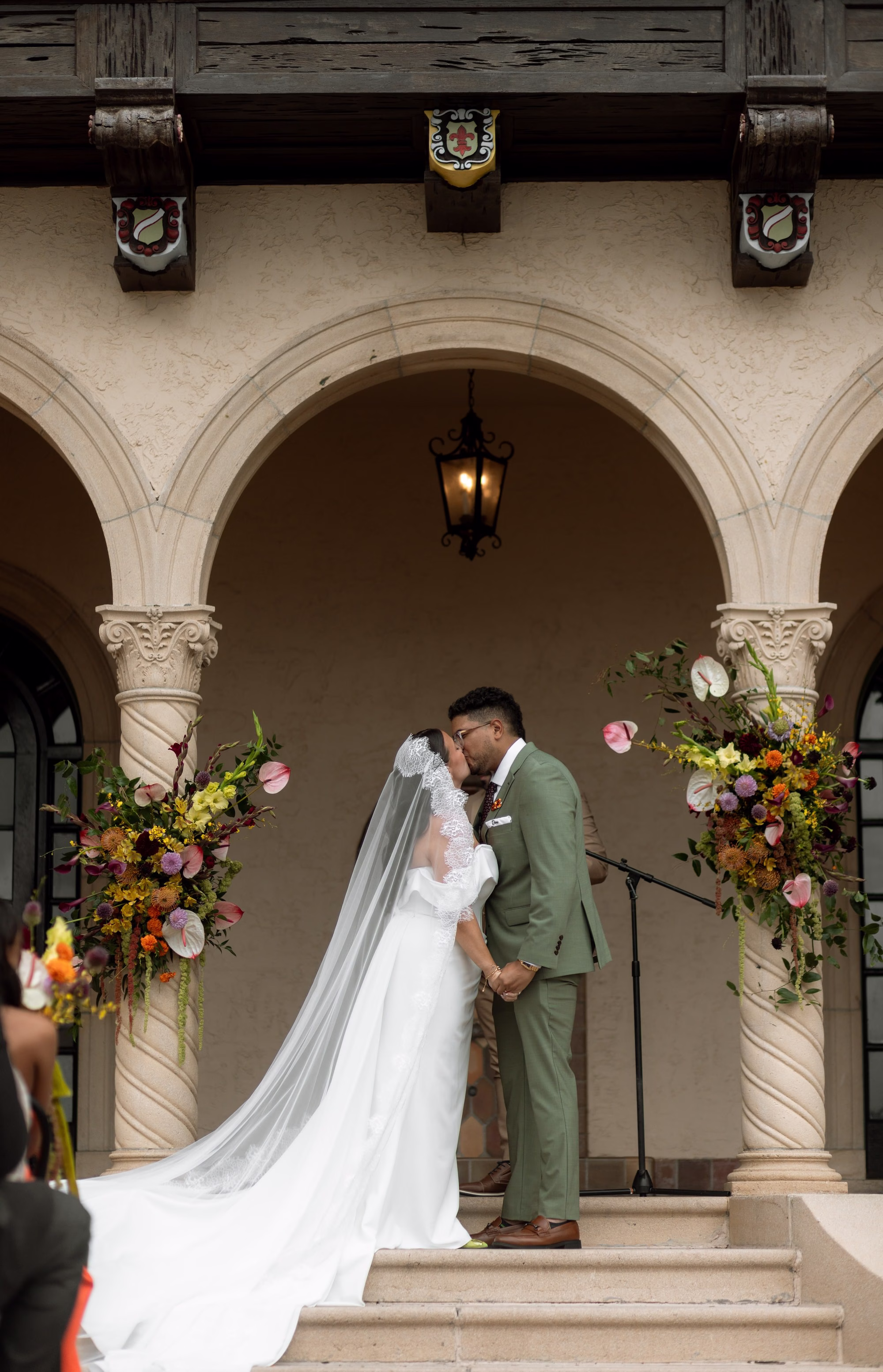 Bride and Groom Wedding Ceremony Just Married Kiss Wedding Portrait | White Off the Shoulder Made with Love Wedding Gown Inspiration | Traditional Lace Cathedral Length Wedding Veil Ideas | Green Groom Suit Inspiration | Tropical Fall Colors Yellow, Orange, Purple, Pink, Red Florals with Greenery Wedding Ceremony Altar Decor Inspiration | Sarasota Event Venue Powel Crosley Estate