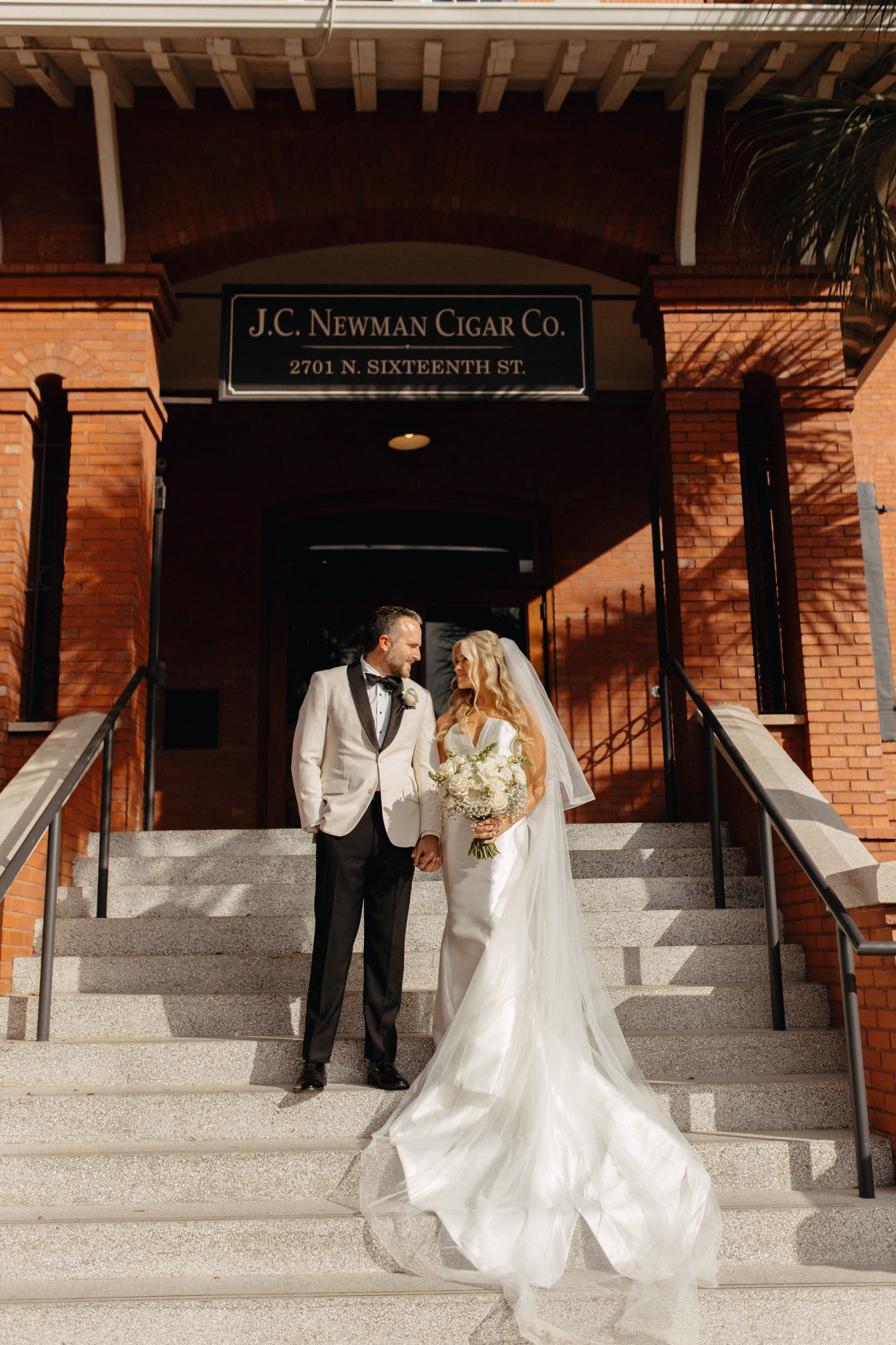 Bride and Groom Wedding Portrait on steps of Ybor Event Venue J.C. Newman Cigar Company | Groom White Tuxedo Jacket Inspiration | Traditional Cathedral Length Chiffon Veil Ideas | Monochromatic White Roses Bridal Bouquet Inspiration | Clearwater Florist Lemon Drops Wedding and Events