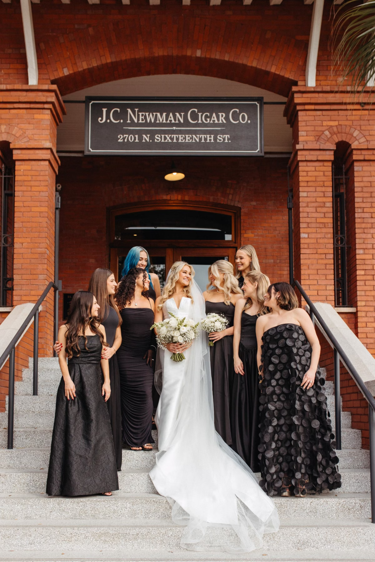 Bride and Bridesmaids on steps of Ybor Event Venue J.C. Newman Cigar Company | Monochromatic White Roses Bridal Bouquet Inspiration | Different Styled Black Bridesmaid Dresses Ideas | Clearwater Florist Lemon Drops Wedding and Events
