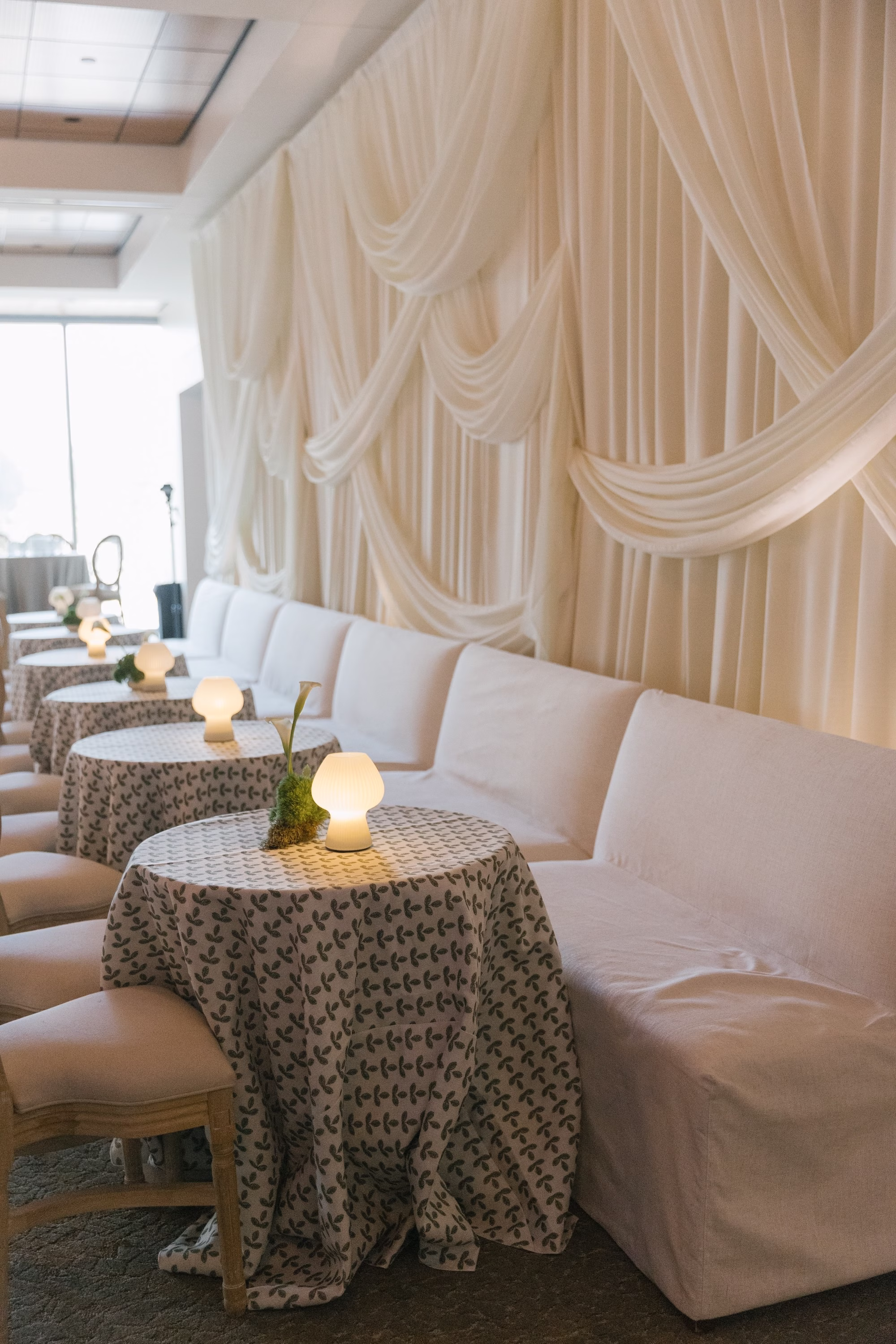 Elegant Ballroom Event Space Wedding Reception | Modern Cream Colored Drapes and Upholstred White Loveseat Chairs Wedding Reception Decor Inspiration | Minimalistic White and Green Centerpiece Floral Arrangements Ideas | Simple Table Lamps on Delicate Floral Linens Decor Inspiration | St Pete Planner Stelloccasions Weddings and Events | Clearwater Kate Ryan Event Rentals