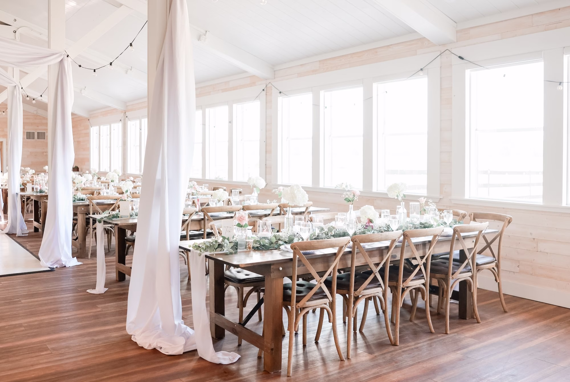 Rustic Elegant White and Green Spring Wedding Wedding Reception Decor Inspiration | Long Wooden Feasting Tables with Greenery Garland Tablescape Decor Ideas | White Draping and Romantic String Lighting Decor Inspiration | Tampa Bay Event Venue Legacy Lane Weddings