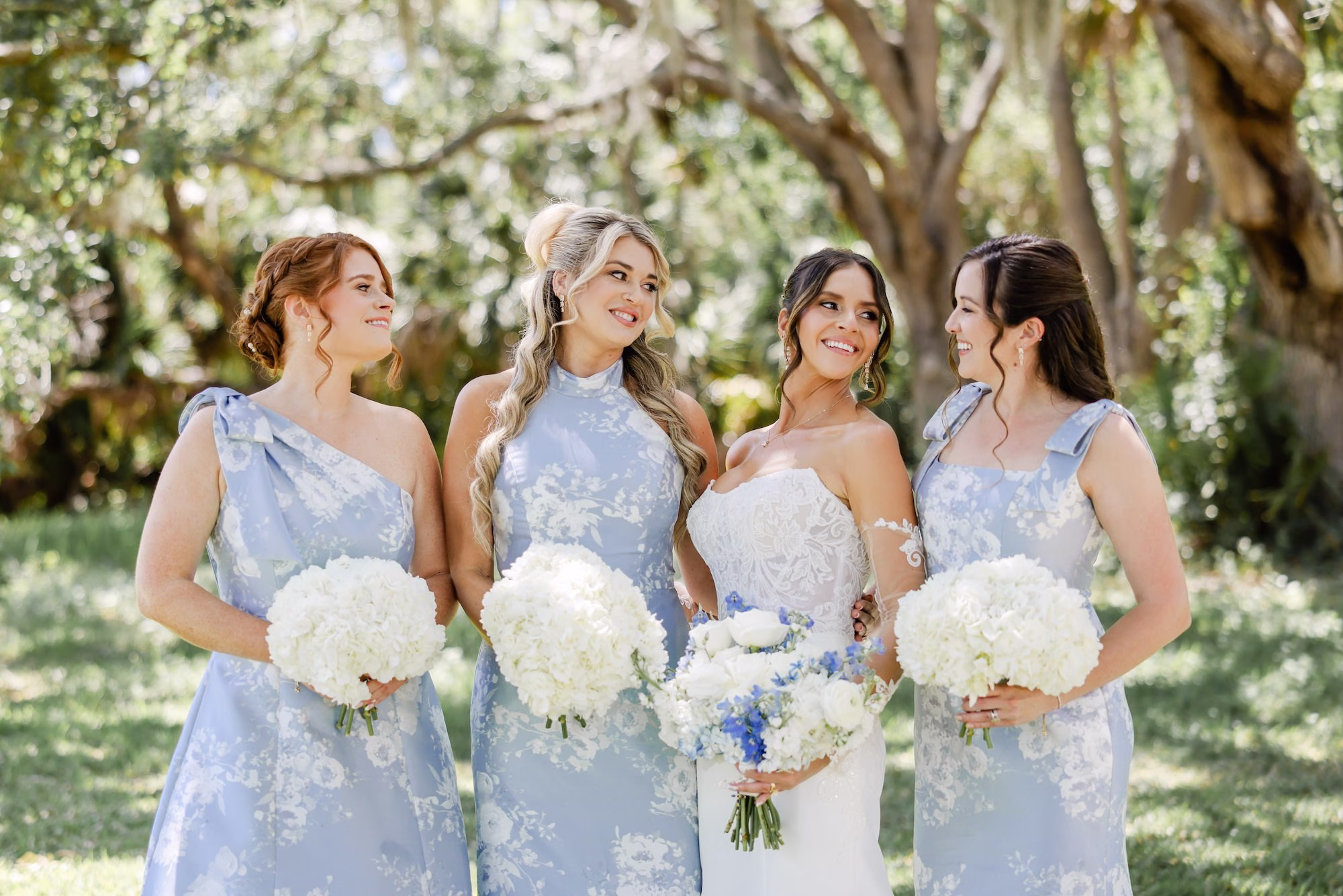 Bride and Bridesmaid Outdoor Spring Wedding Portrait | Blue and White Floral Mismatched Bridesmaid Dress Styles Inspiration | Elegant Strapless The Dessy Group White Wedding Gown with Lace Bodice and Sleeves Ideas | Lush White Roses and Hydrangeas with Blue Delphinium Bridal Bouquet Inspiration | Monochromatic White Hydrangeas Bridesmaid Bouquet Ideas | Full Glam Bridal Makeup with Soft Curled Half Up Half Down Hair Inspiration | Clearwater Photographer Lifelong Photography Studio | Tampa Boutique Truly Forever Bridal | Ybor Hair and Makeup Artist Adore Bridal Hair and Makeup | Content Creator Behind The Vows