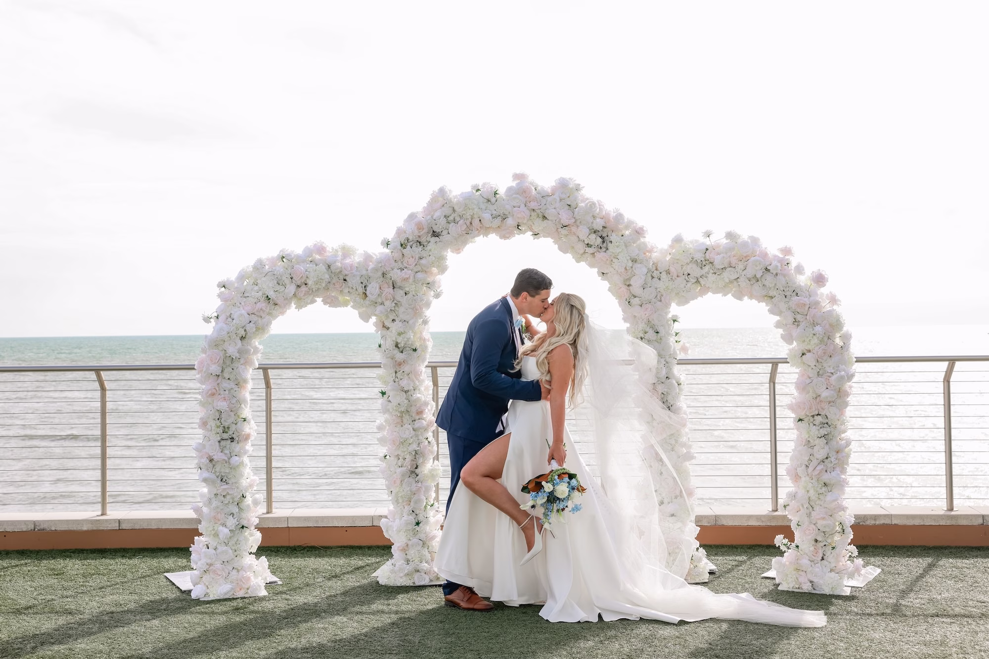 23Elegant Light Blue Coastal Florida Wedding Ideas Clearwater Venue Opal Sands - Elegant Blue Modern Coastal Wedding | Opal Sands Clearwater Beach Bride and Groom Kissing Under Triple White Floral Arch Overlooking the Gulf Wedding Portrait | Rooftop Lawn Wedding Ceremony at Clearwater Beach Venue Opal Sands Resort | Photography Lifelong Photography Studio