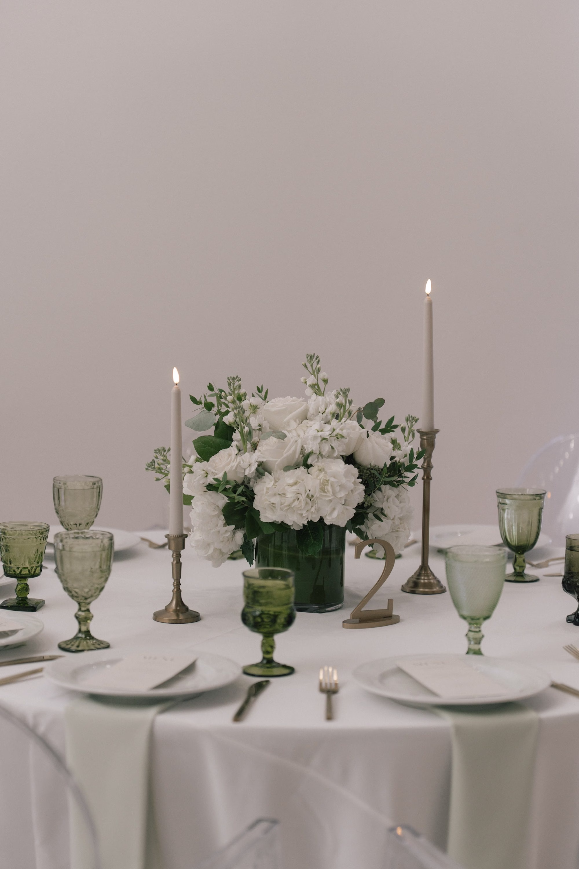 Lush White Floral Centerpiece of Roses and Hydrangeas Decor Inspiration | White Linen with Metal Candle Holders and White Taper Candles Tablescape Decor Ideas | Mismatched Green Depression Glass Stemware Inspiration | Wedding Treasure Island Caterer Amici's Catered Cuisine