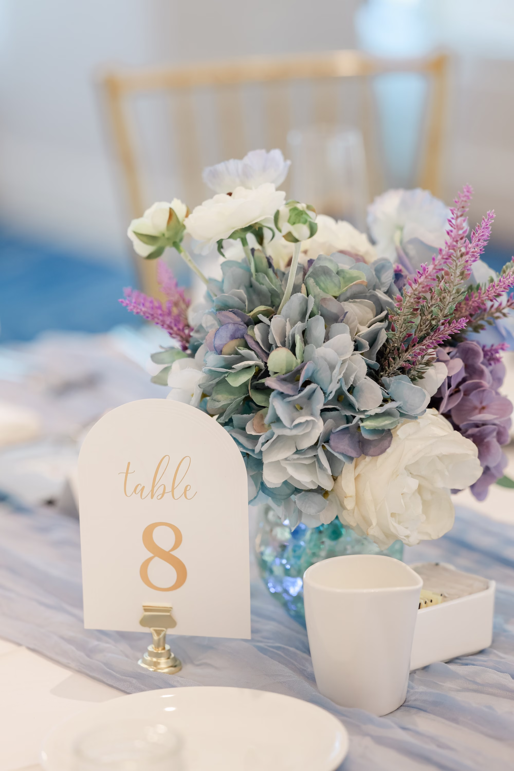 Romantic Ocean Blue Wedding Reception Inspiration | Lush Floral Arrangement of Vibrant Blue Hydrangeas, White Roses and Purple Lavender Floral Tablescape Centerpiece Decor Ideas | Elegant Arched Gold and White Table Number Reception Decor Ideas