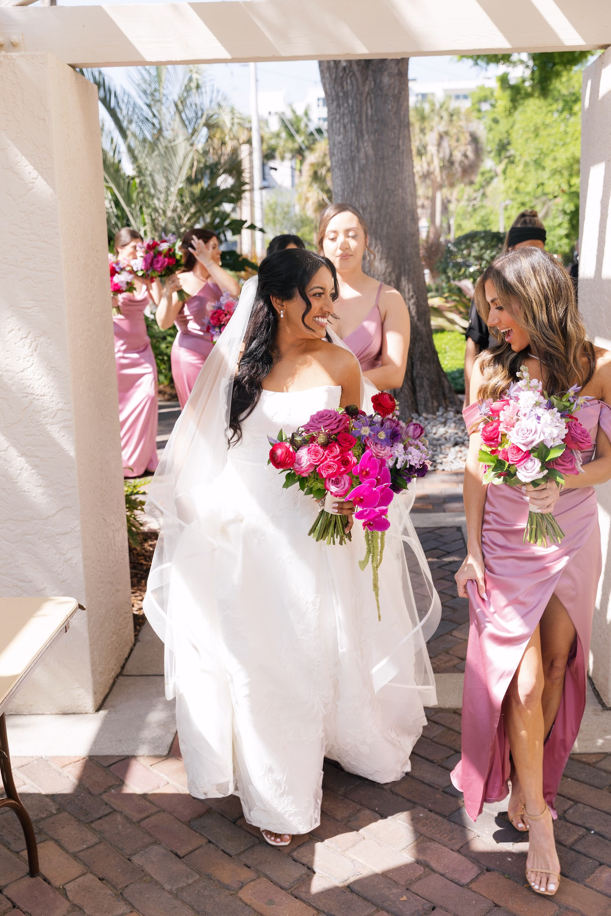 Bride in White Strapless Ballgown and Cathedral Veil with Vibrant Pink and Purple Orchid Bouquet Walking with Bridesmaids in Dusty Rose Satin Dresses | St. Pete Florist Bruce Wayne Florals