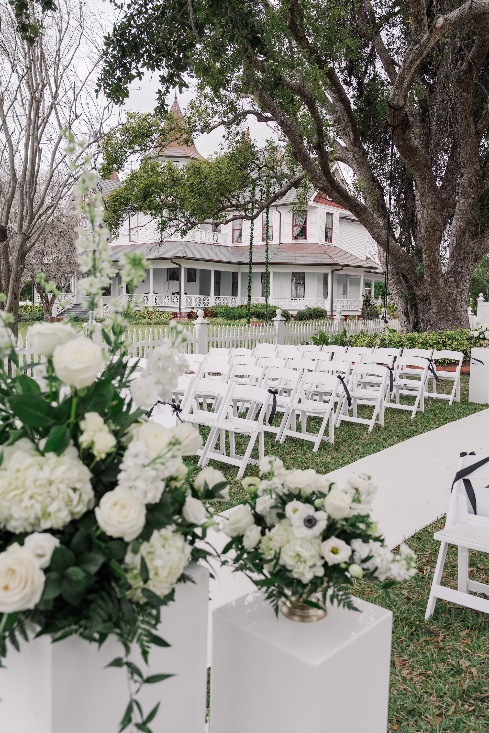 White Rose and Greenery Aisle Arrangements Lining White Aisle Runner with White Folding Chairs and Old Florida Home Wedding Ceremony Ideas | Palmetto Venue Palmetto Riverside Bed and Breakfast | Rentals Kate Ryan Event Rentals