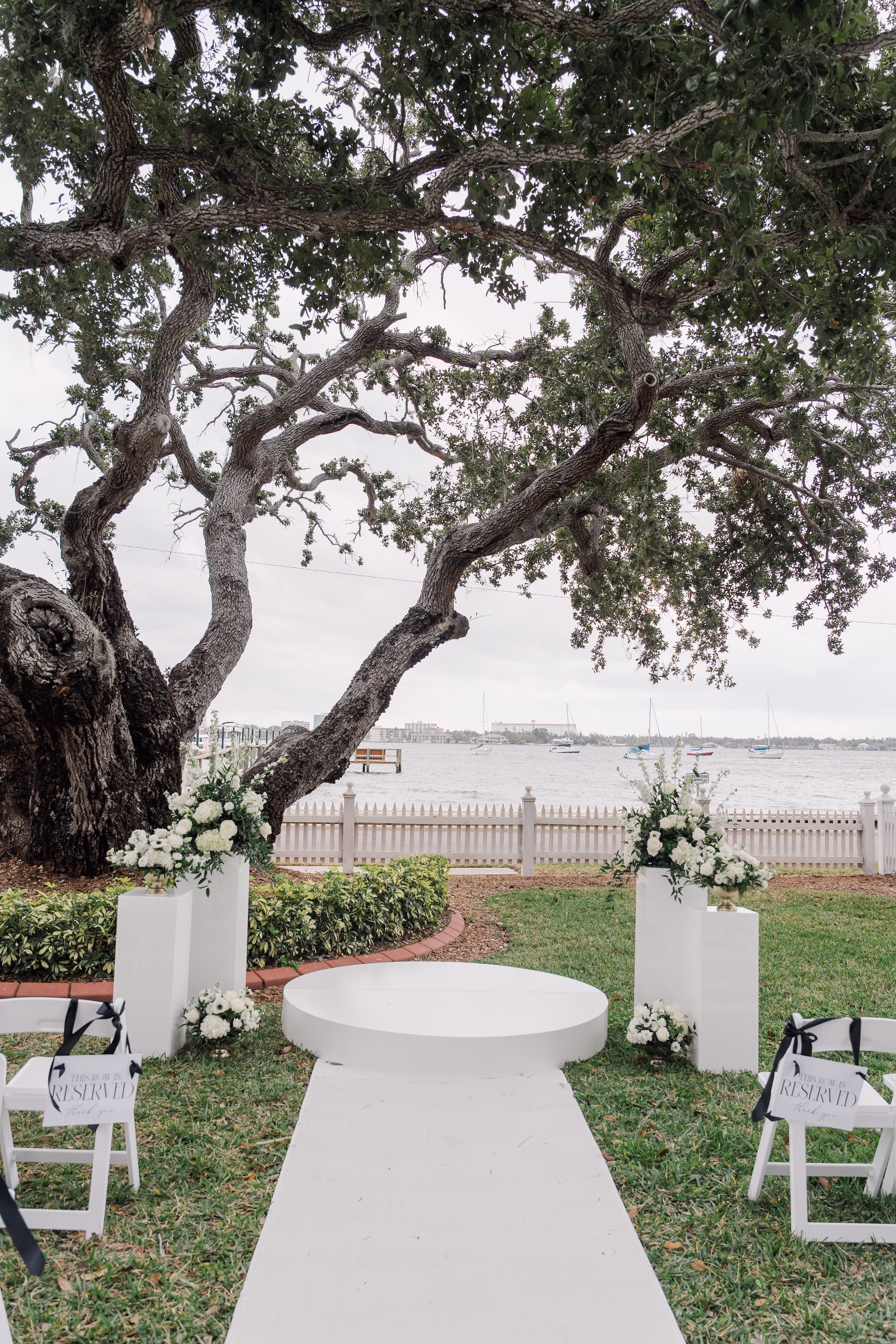 Riverfront Wedding Ceremony Setup with Round White Platform, White Aisle Runner and Pedestal Florals Under Live Oak Ideas | Palmetto Venue Palmetto Riverside Bed and Breakfast | Rentals Kate Ryan Event Rentals