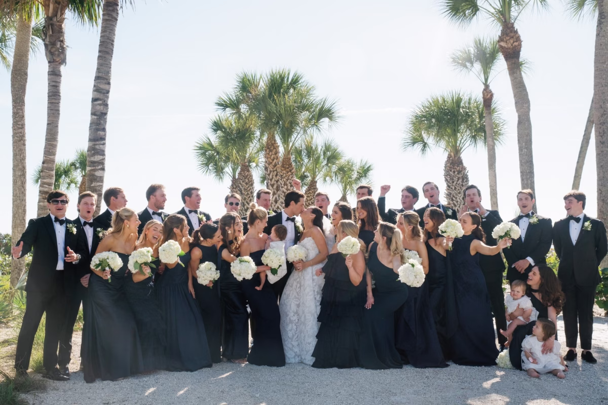 24Coastal Nancy Meyers Inspired Florida Beach Wedding - Coastal Blue, White, and Green Sarasota Wedding | The Resort at Longboat Key Club Wedding Party Portrait on the Beach Under Palm Trees in Black Tuxedos and Navy Blue Bridesmaid Dresses with White Bouquets