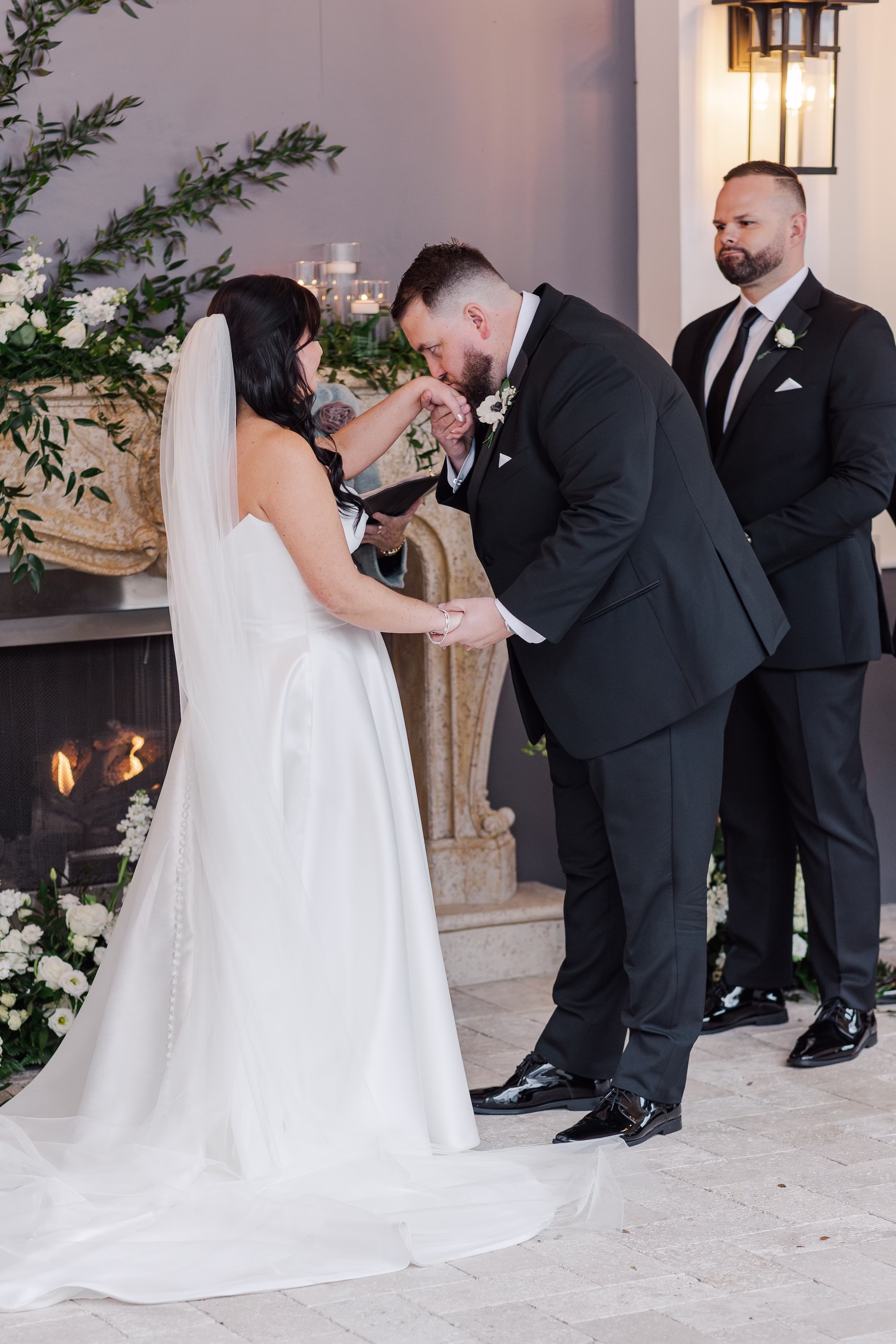 Groom Kissing Bride's Hand Wedding Ceremony Portrait in Front of Greenery-Draped Fireplace