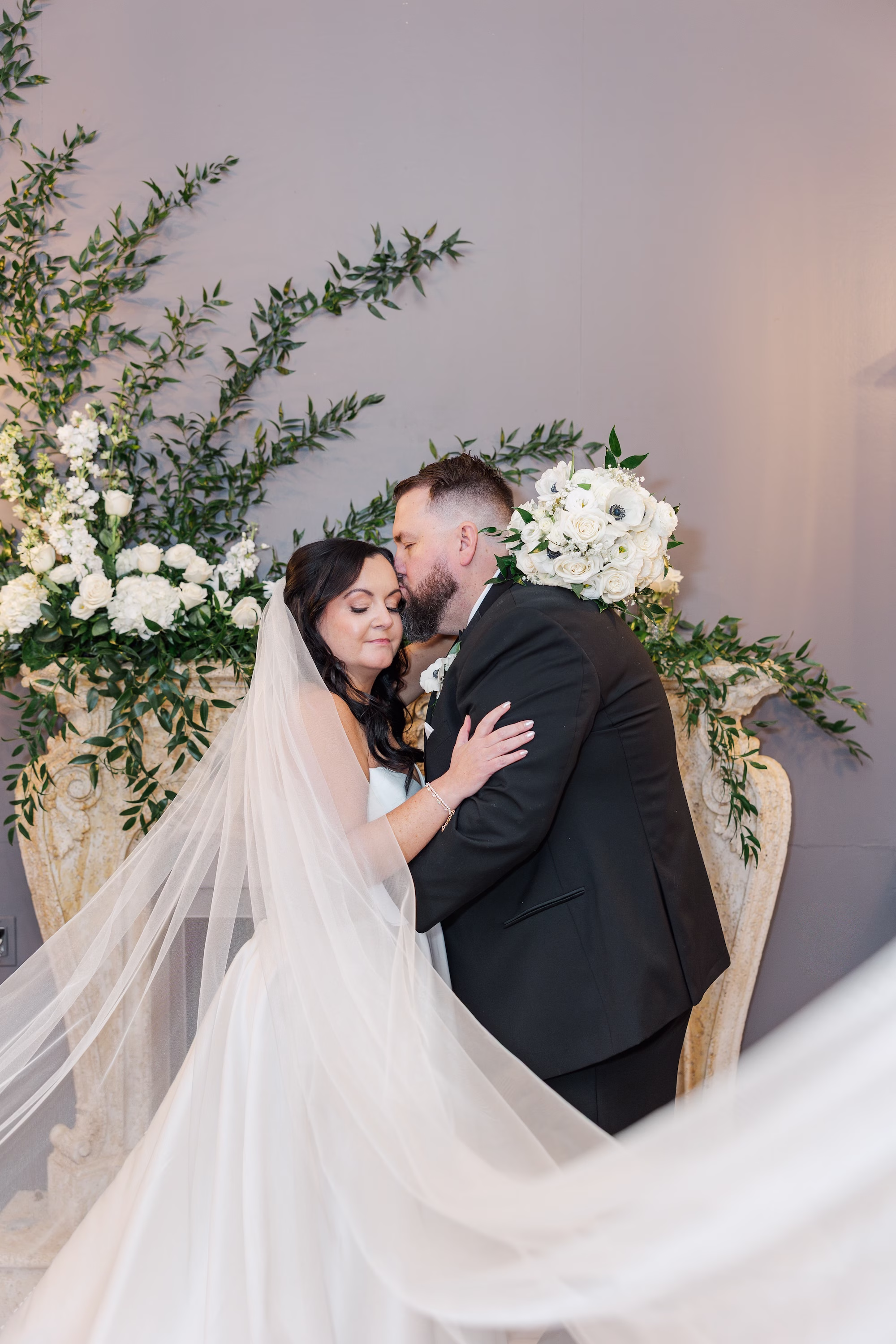 Bride and Groom Kiss Wedding Portrait with Cathedral Veil in Front of White Rose and Greenery Fireplace Installation