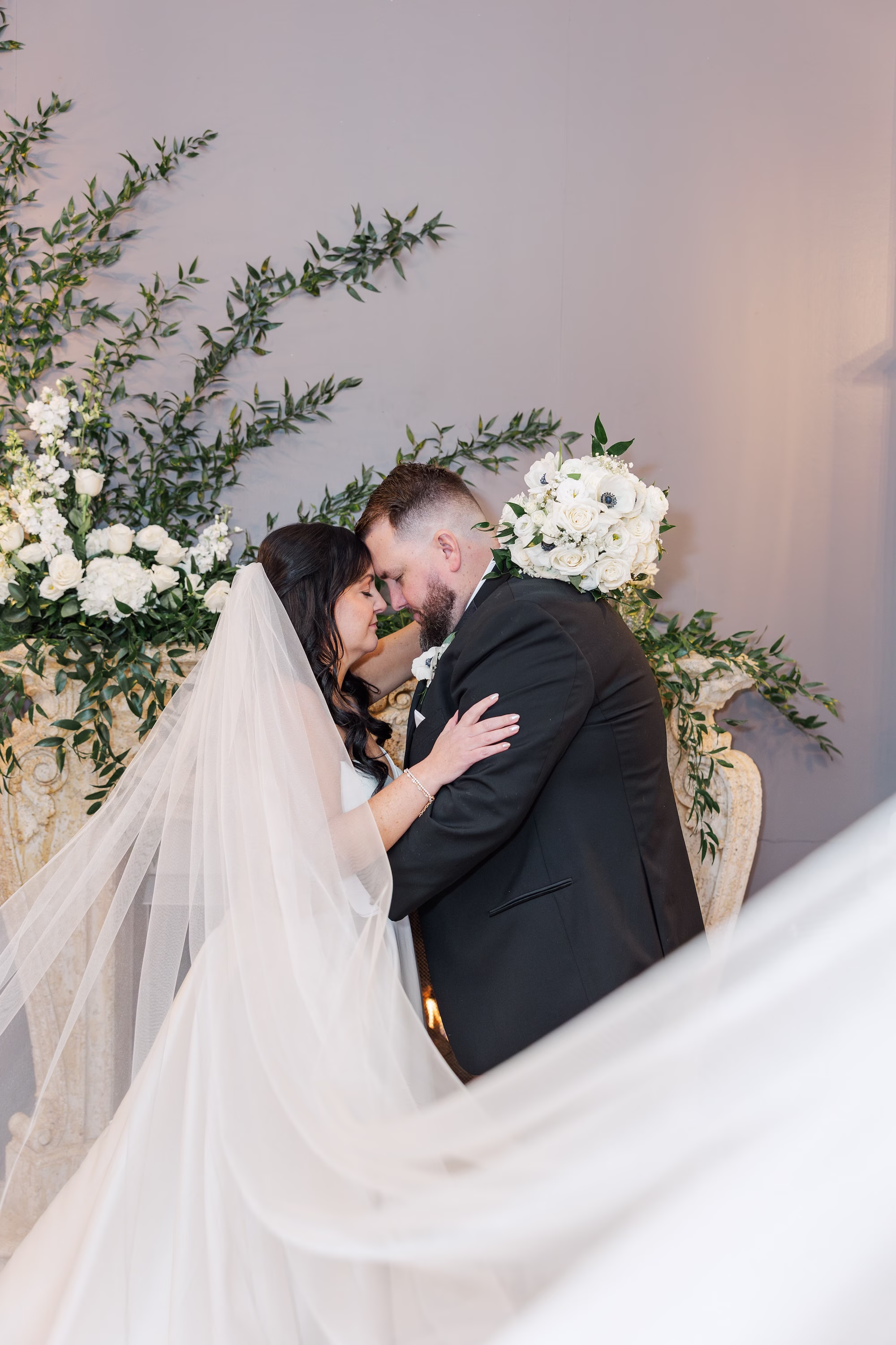 Bride and Groom Forehead Kiss Wedding Portrait with Cathedral Veil and White Rose Greenery Backdrop