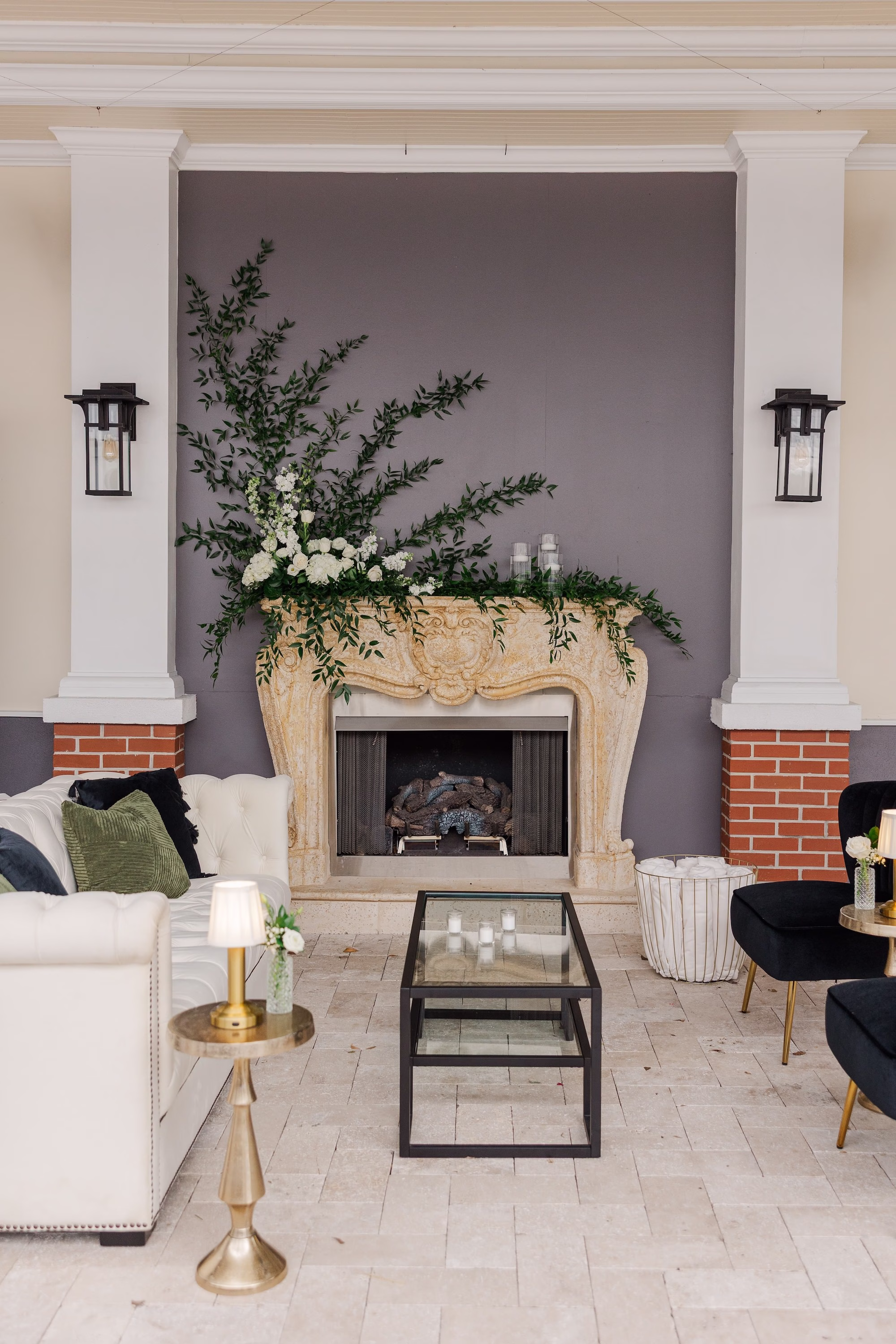 White Tufted Couch and Black Velvet Chairs Lounge Area in Front of Greenery-Draped Stone Fireplace Wedding Cocktail Hour Lounge Ideas | Palmetto Rentals Kate Ryan Event Rentals