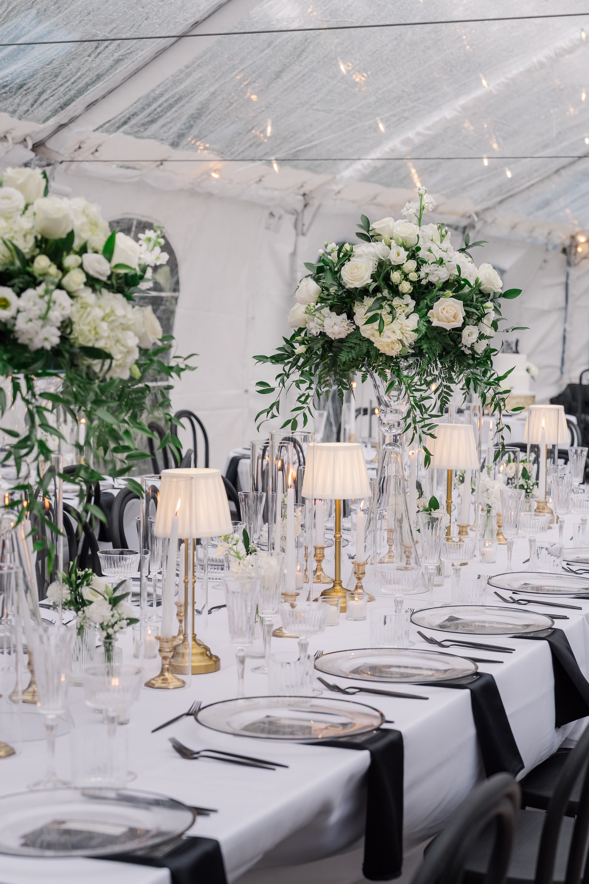 Long White Wedding Reception Table with Tall White Rose Centerpieces, Gold Lamp Shade Candlesticks, Black Napkins and Black Bentwood Chairs Ideas | Palmetto Rentals Kate Ryan Event Rentals
