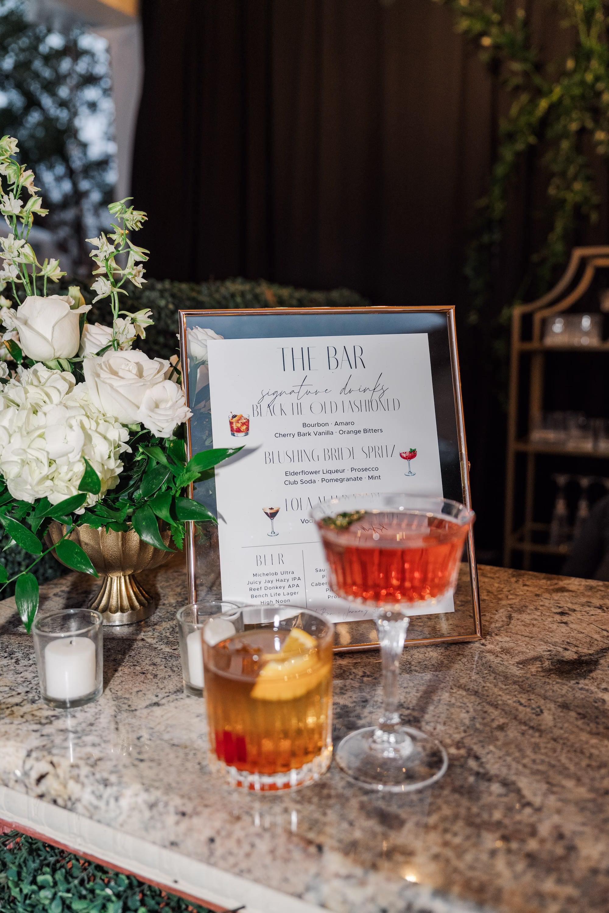 The Bar Signature Drinks Sign with Old Fashioned and Aperol Spritz Cocktails and White Rose Arrangement Wedding Bar Ideas