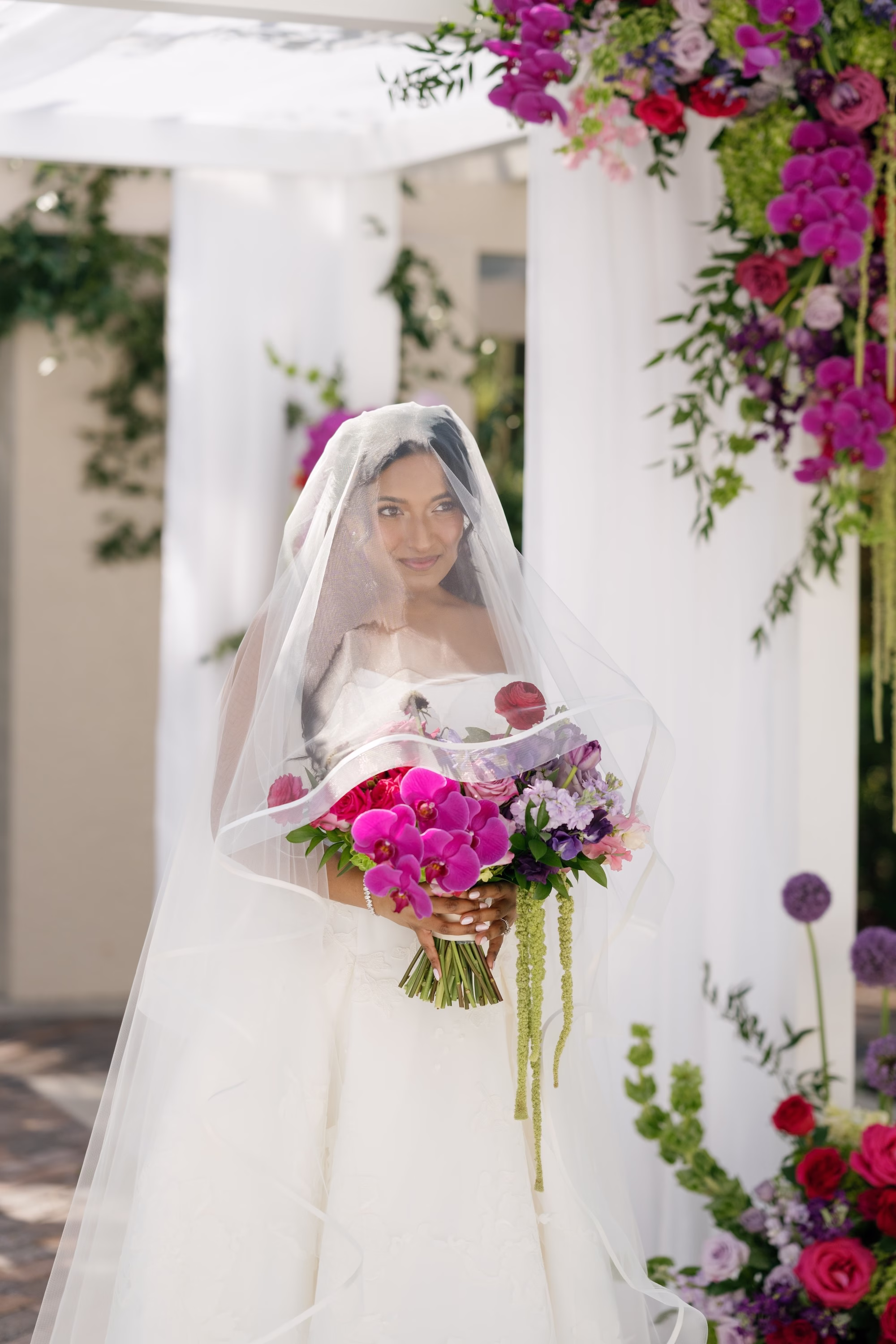 Bride in Blusher Veil Holding Vibrant Pink and Purple Bouquet Under Floral-Draped Chuppah with Fuchsia Orchid Arrangement | Jewish Wedding | St. Pete Florist Bruce Wayne Florals
