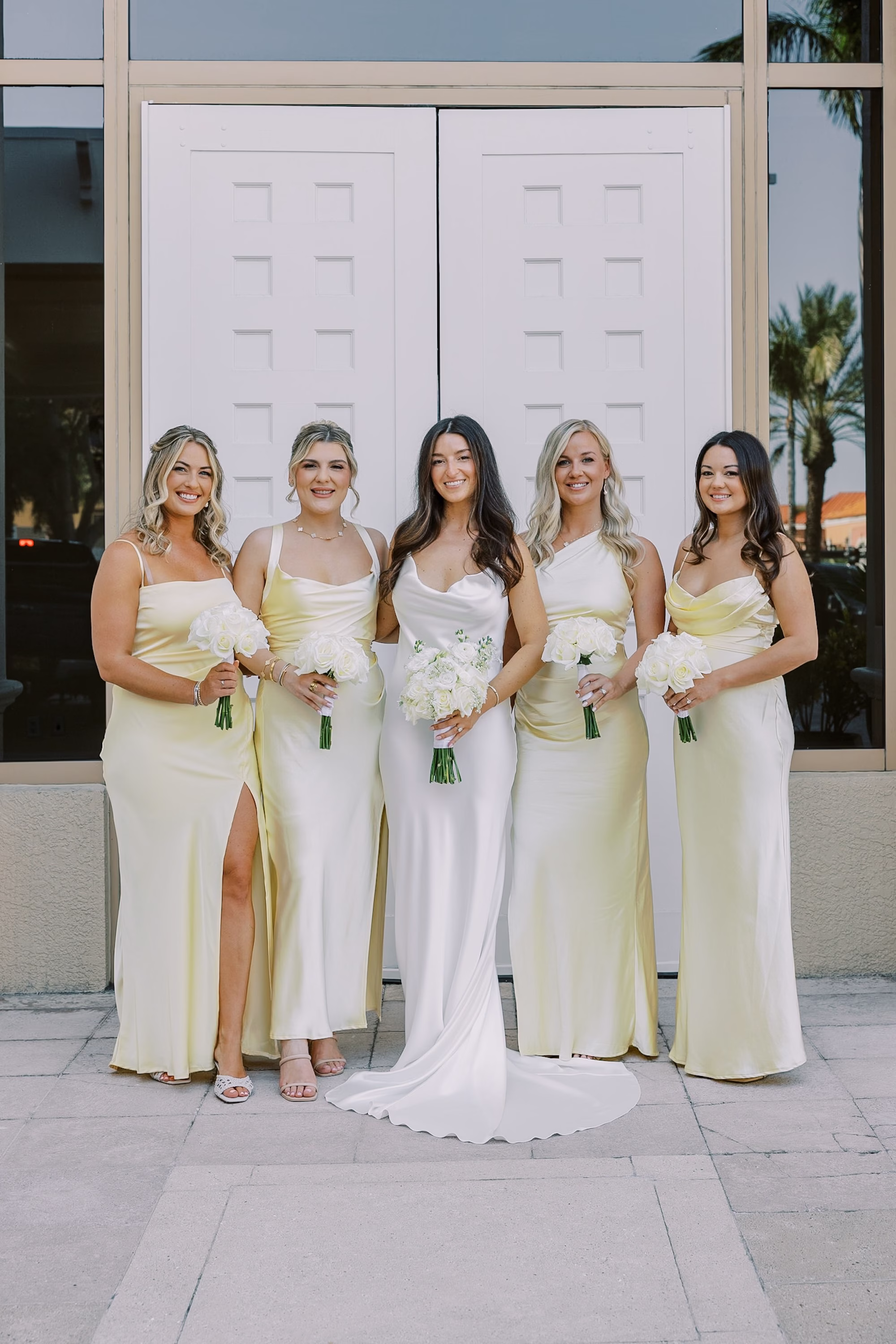 Bride with Bridesmaids in Mismatched Pale Yellow Satin Dresses Holding White Bouquets in Front of White Double Doors