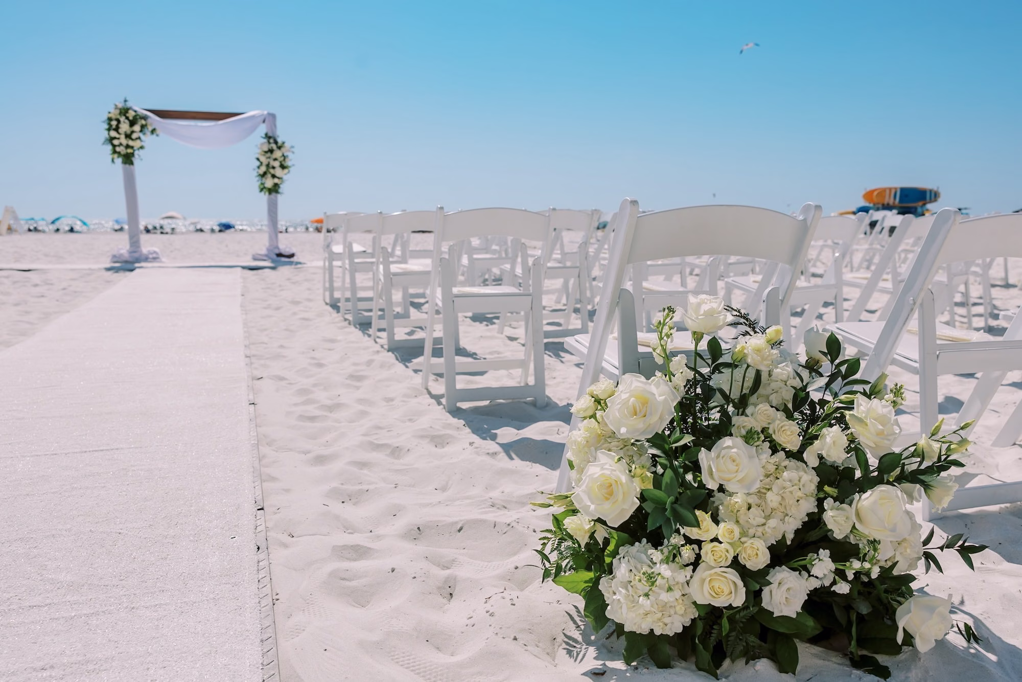 Beach Wedding Ceremony Aisle with White Folding Chairs, White Rose Arrangements and White Draped Arch on the Sand Ideas | Beach Wedding Ceremony | Clearwater Planner Special Moments Event Planning | Rentals Gabro Event Services