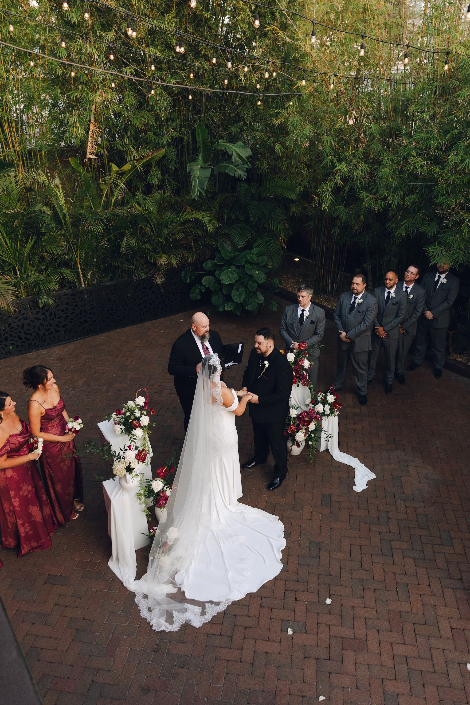 16Burgundy Vintage Romance Wedding Ideas St Pete Venue NOVA 535 - Vintage Romance Pink and Burgundy Downtown St. Pete Wedding | NOVA 535 Aerial View of Bride and Groom Exchanging Vows Under String Lights at Bamboo Courtyard Wedding Ceremony | Courtyard Wedding Ceremony at St. Pete Venue NOVA 535