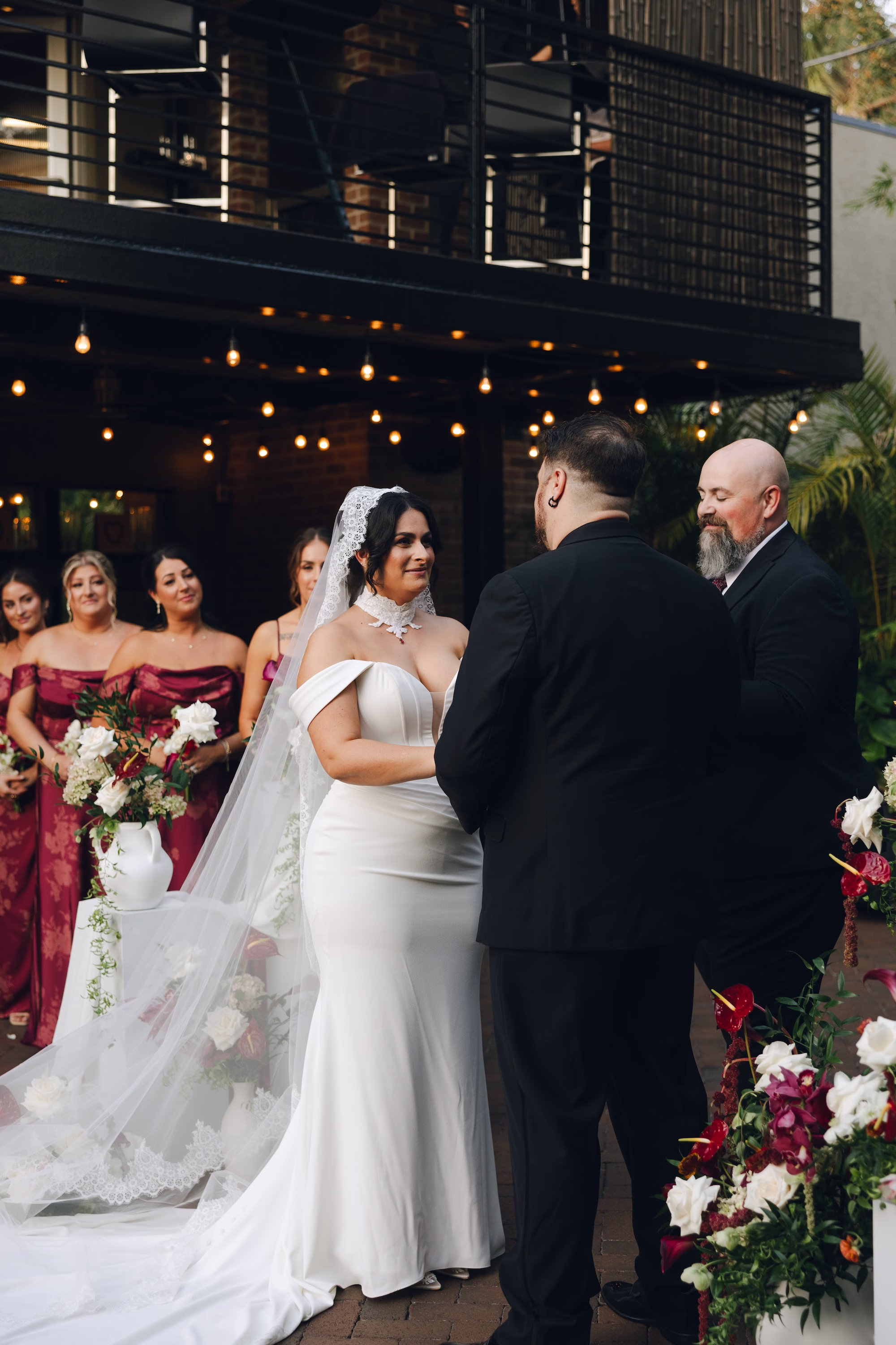 18Burgundy Vintage Romance Wedding Ideas - Vintage Romance Pink and Burgundy Downtown St. Pete Wedding | NOVA 535 Bride and Groom Exchanging Vows Beside Burgundy Anthurium and White Rose Ceremony Arrangement with Bridesmaids in Burgundy | Courtyard Wedding Ceremony
