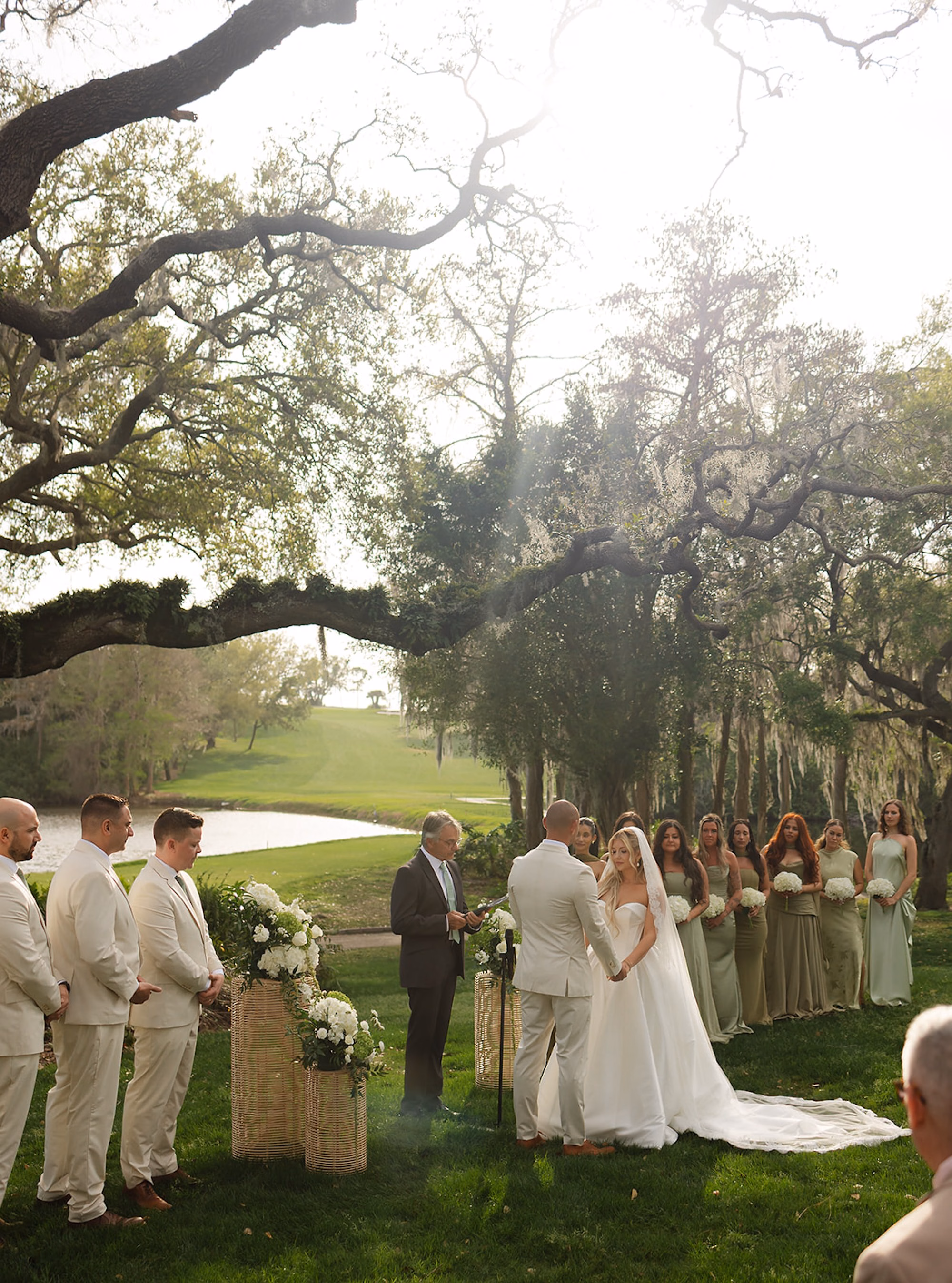 Bride and Groom Exchanging Vows Under Live Oak with Wedding Party in Sage Green and Cream Overlooking Golf Course | Lakeside Lawn Wedding Ceremony at Palm Harbor Venue Innisbrook Golf & Spa Resort
