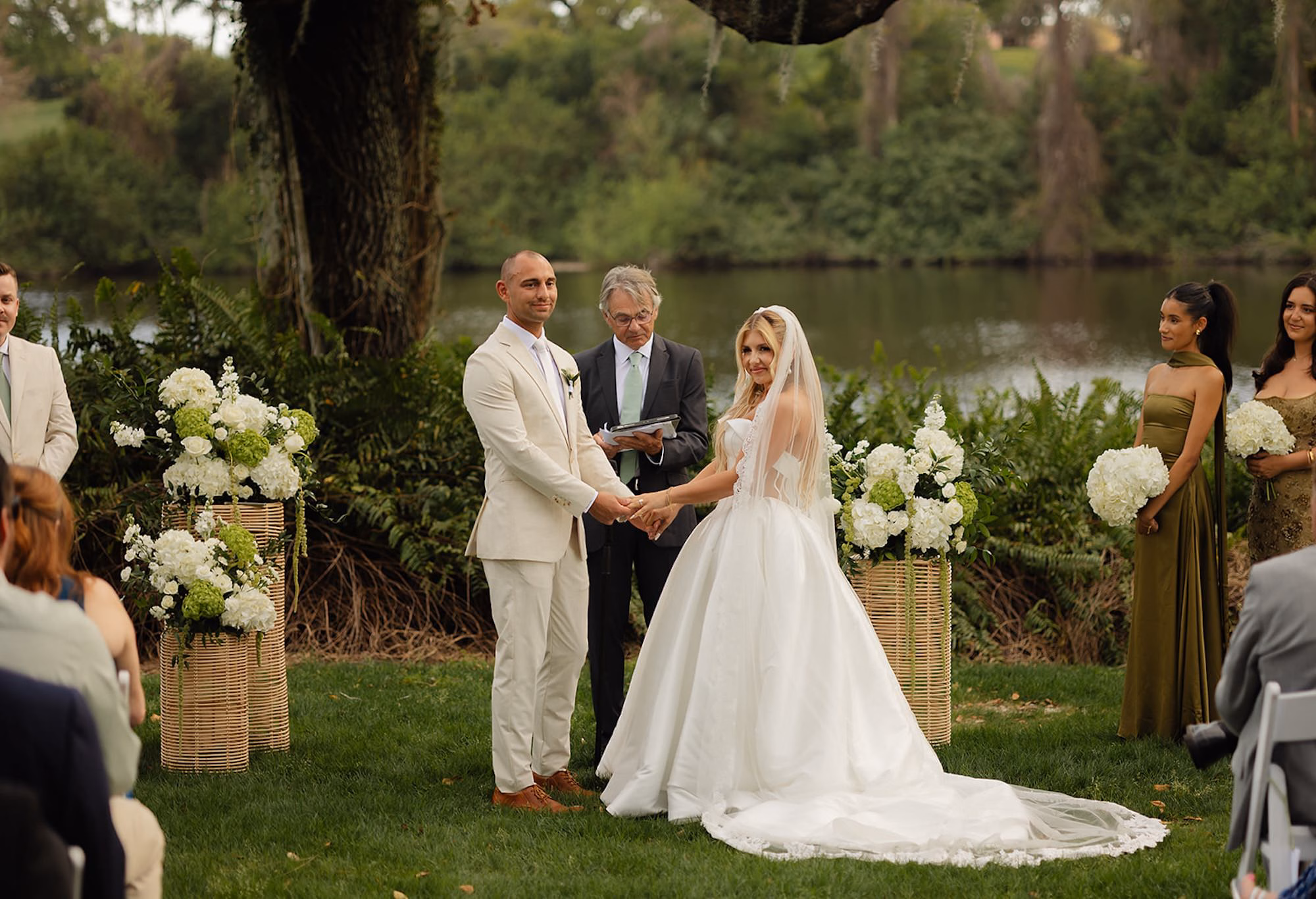 Bride and Groom Holding Hands During Vows Beside White Hydrangea Pedestal Arrangements with Lake Behind | Lakeside Lawn Wedding Ceremony at Palm Harbor Venue Innisbrook Golf & Spa Resort