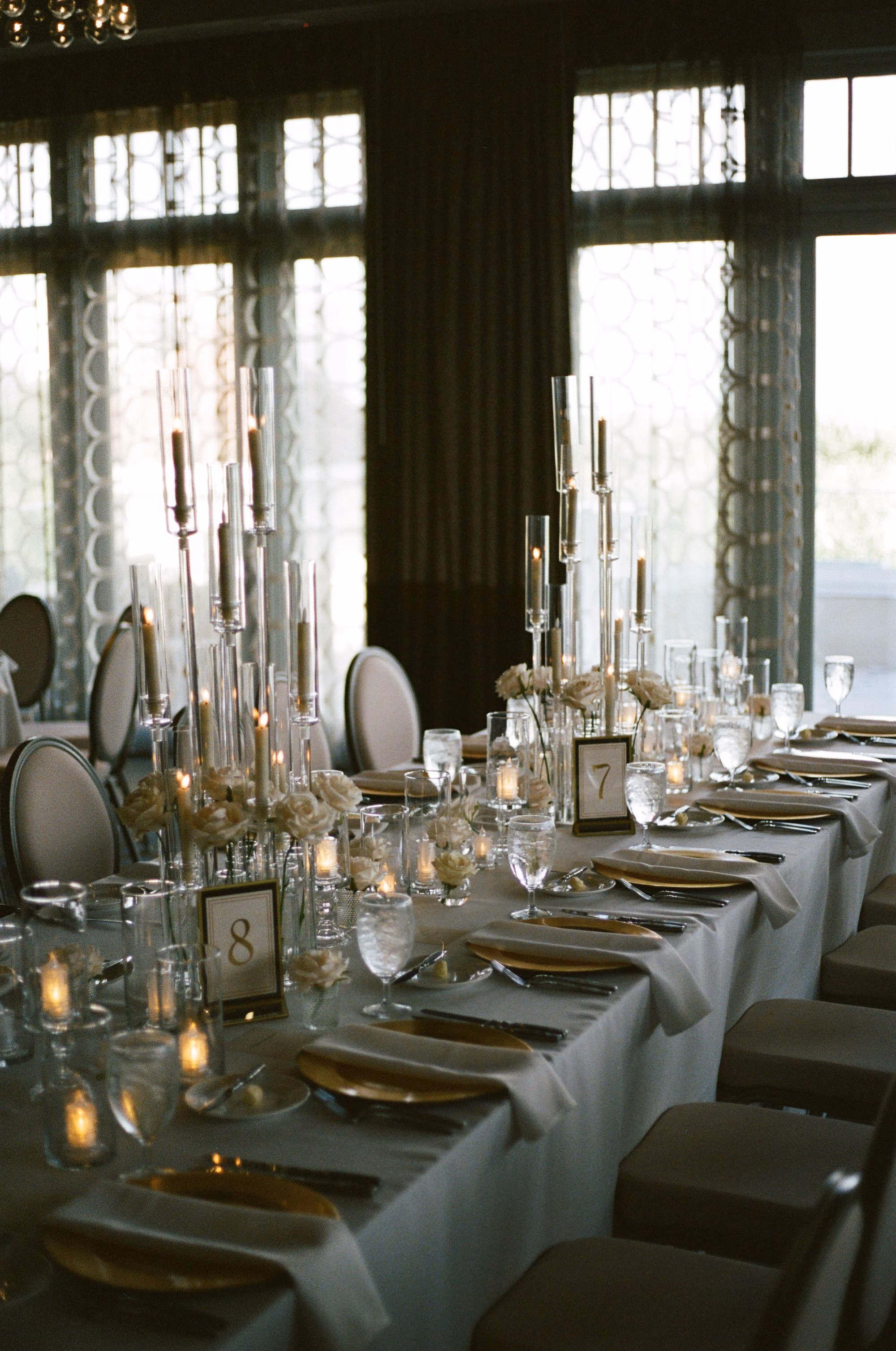 Long Wedding Reception Table with White Linens, Gold Chargers, Taper Candles and White Floral Centerpieces by Arched Windows Decor Ideas