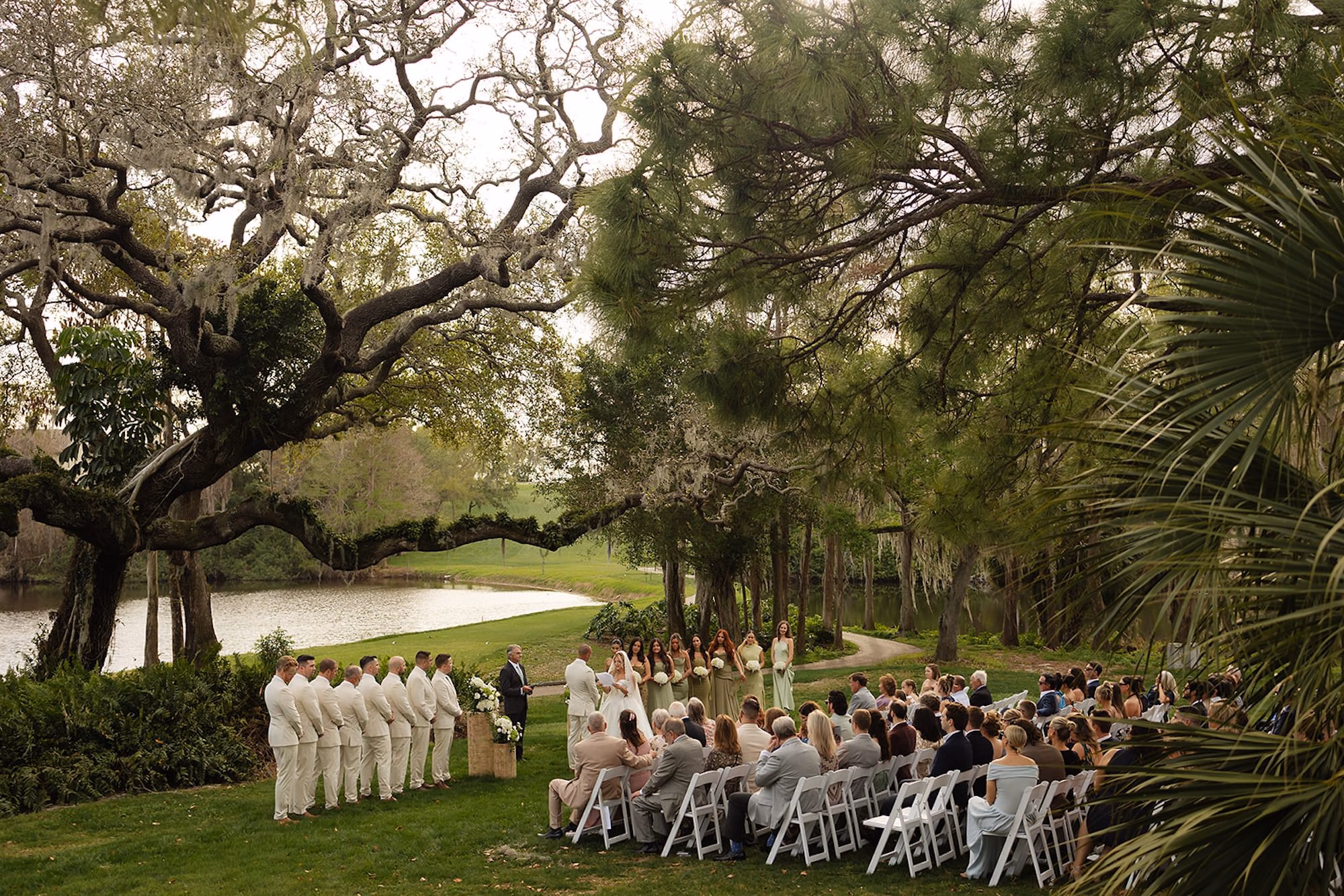 Wide View of Lakeside Wedding Ceremony Under Sprawling Oak Tree with Guests Seated in White Folding Chairs | Lakeside Lawn Wedding Ceremony at Palm Harbor Venue Innisbrook Golf & Spa Resort