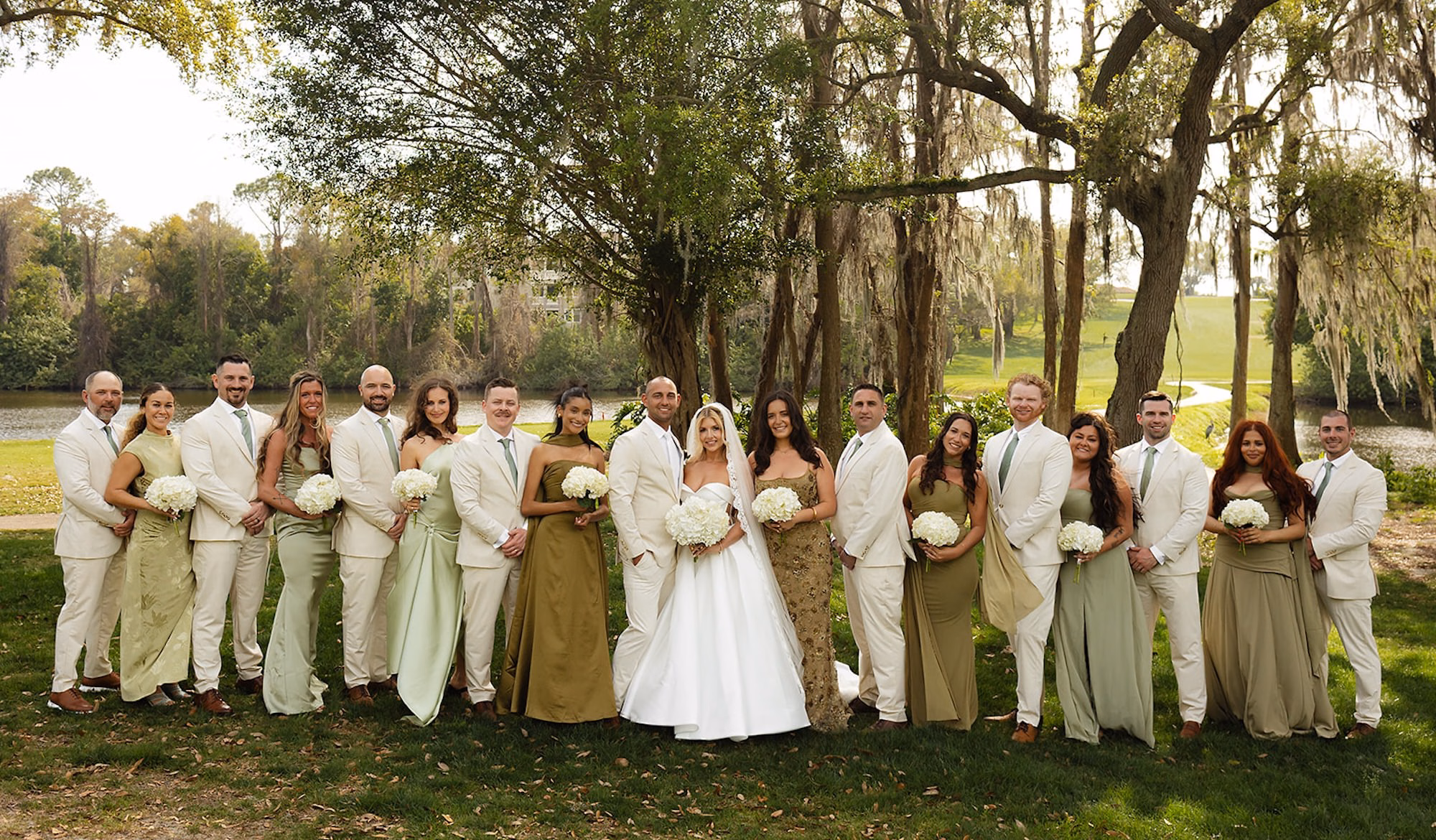 Wedding Party in Sage Green Dresses and Cream Suits with White Hydrangea Bouquets Under Mossy Trees by Lake