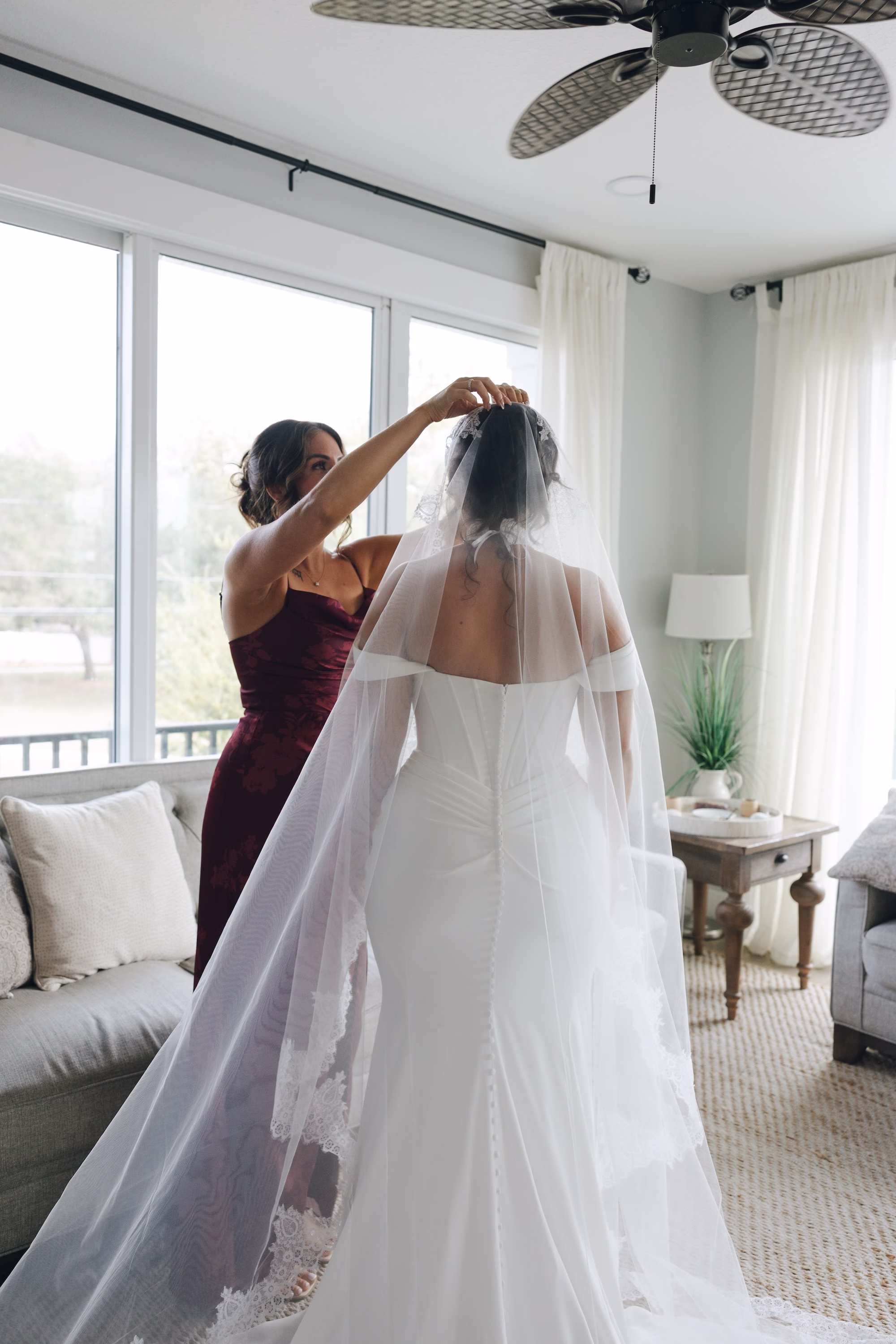 2Burgundy Vintage Romance Wedding Ideas - Vintage Romance Pink and Burgundy Downtown St. Pete Wedding | NOVA 535 Bridesmaid in Burgundy Satin Dress Placing Lace-Trimmed Cathedral Veil on Bride in Off-the-Shoulder White Wedding Dress