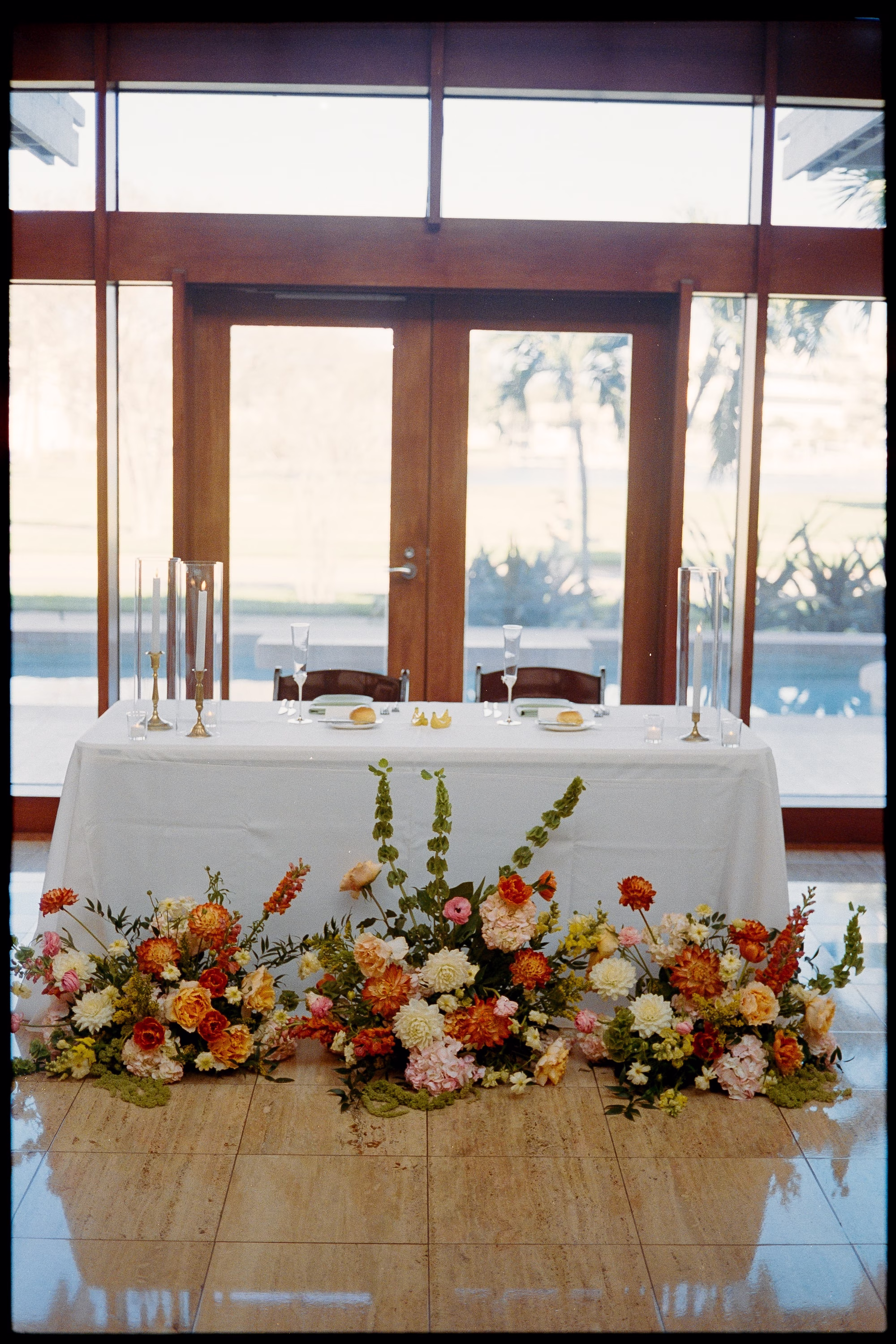 Sweetheart Table with Lush Orange, Peach and White Floral Garland and Taper Candles in Front of Glass Doors Decor Ideas | St. Pete Planner Coastal Coordinating | Florist Monarch Events and Design | Event Venue The Poynter Institute