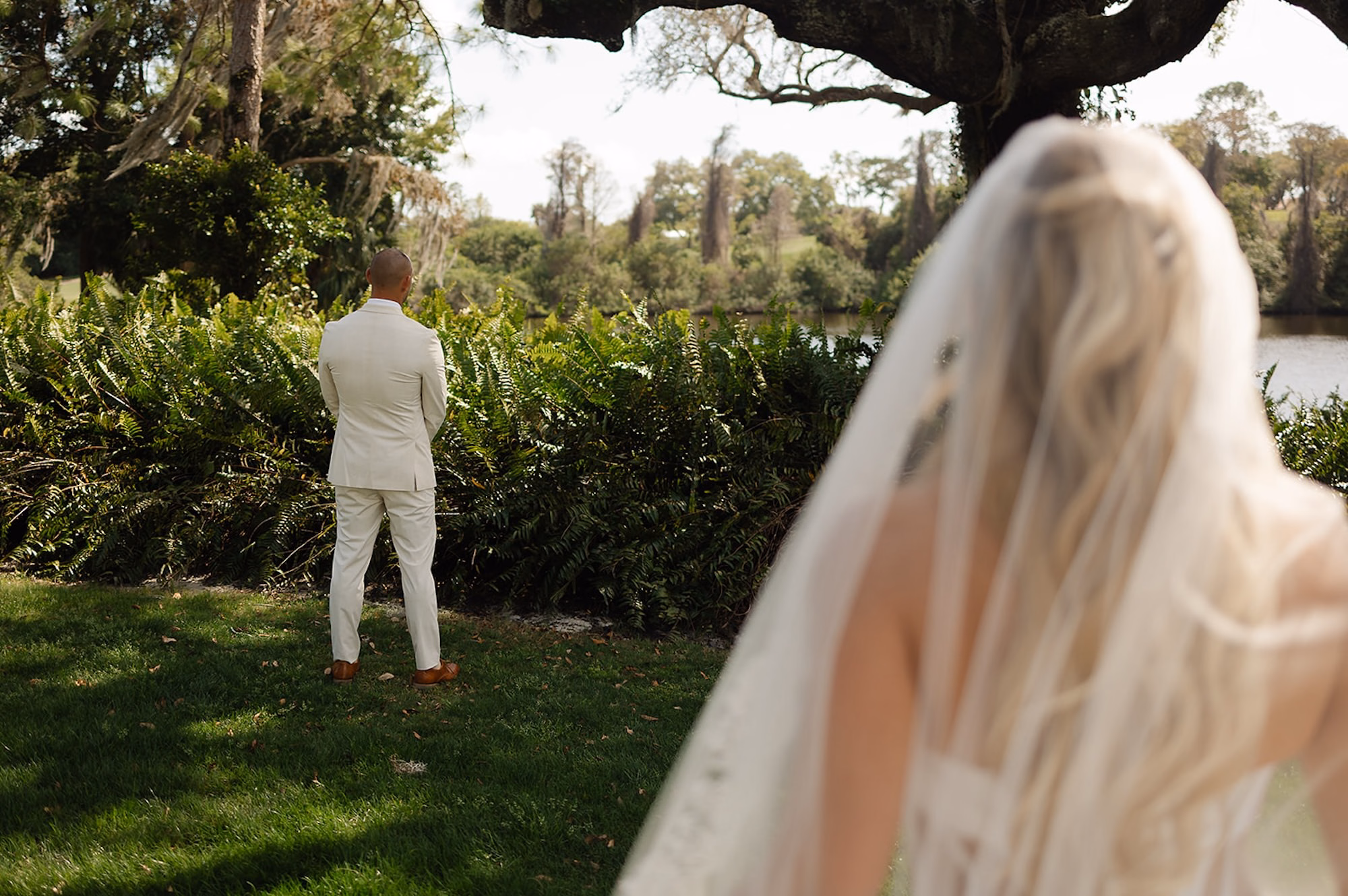 Groom in Cream Linen Suit Waiting for First Look as Bride in Cathedral Veil Approaches on Lawn