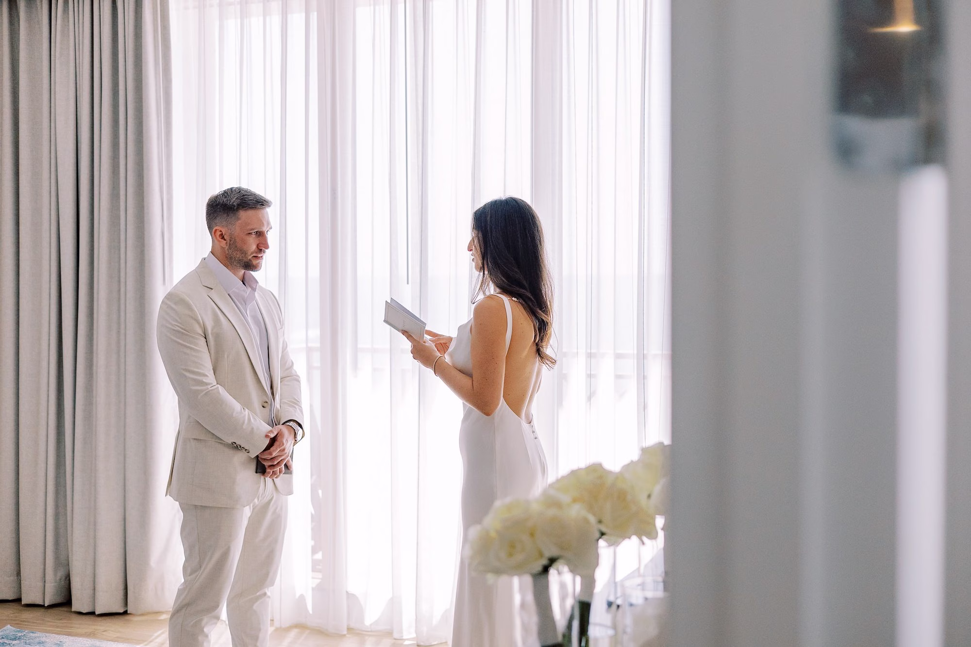 Bride Reading Private Vows to Groom in Tan Suit Beside White Rose Arrangement